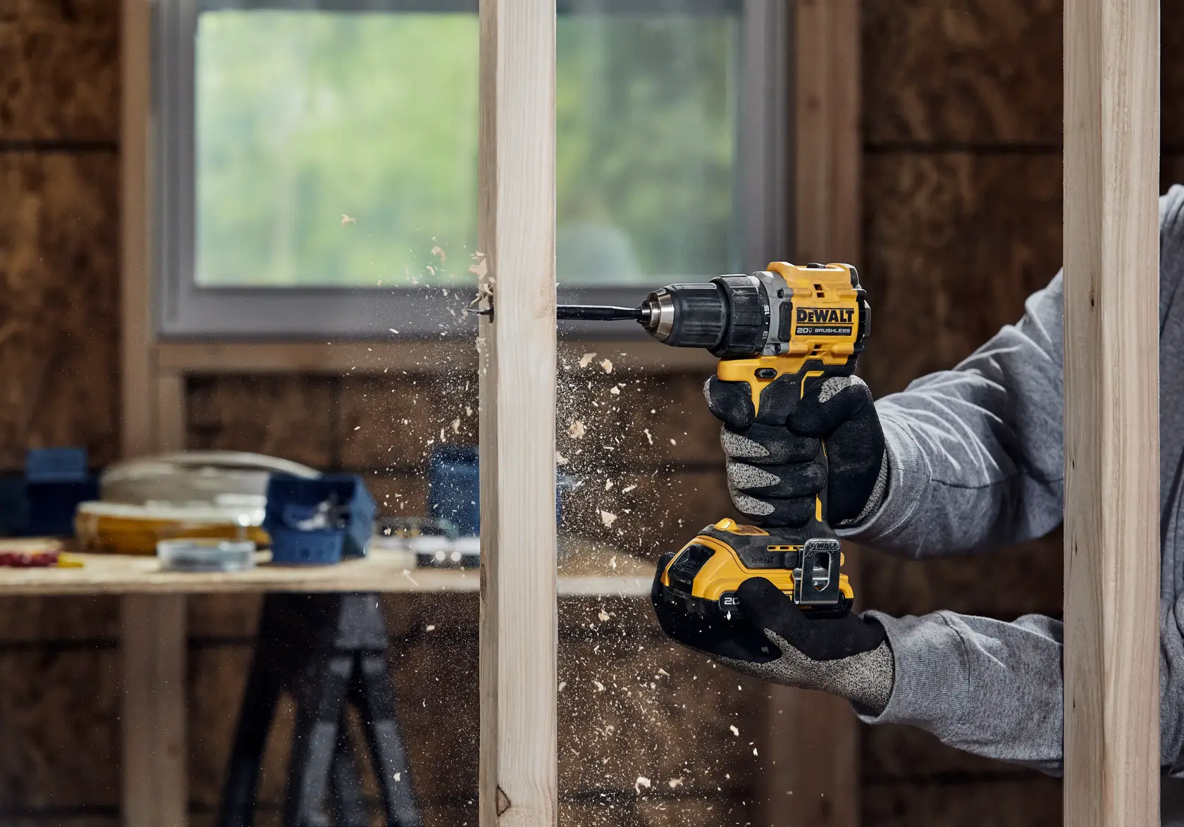 A person using a DEWALT cordless power drill to drill a hole in a vertical wooden beam, with wood shavings flying. The person is wearing gloves and a sweatshirt. The setting appears to be a workshop or construction area.