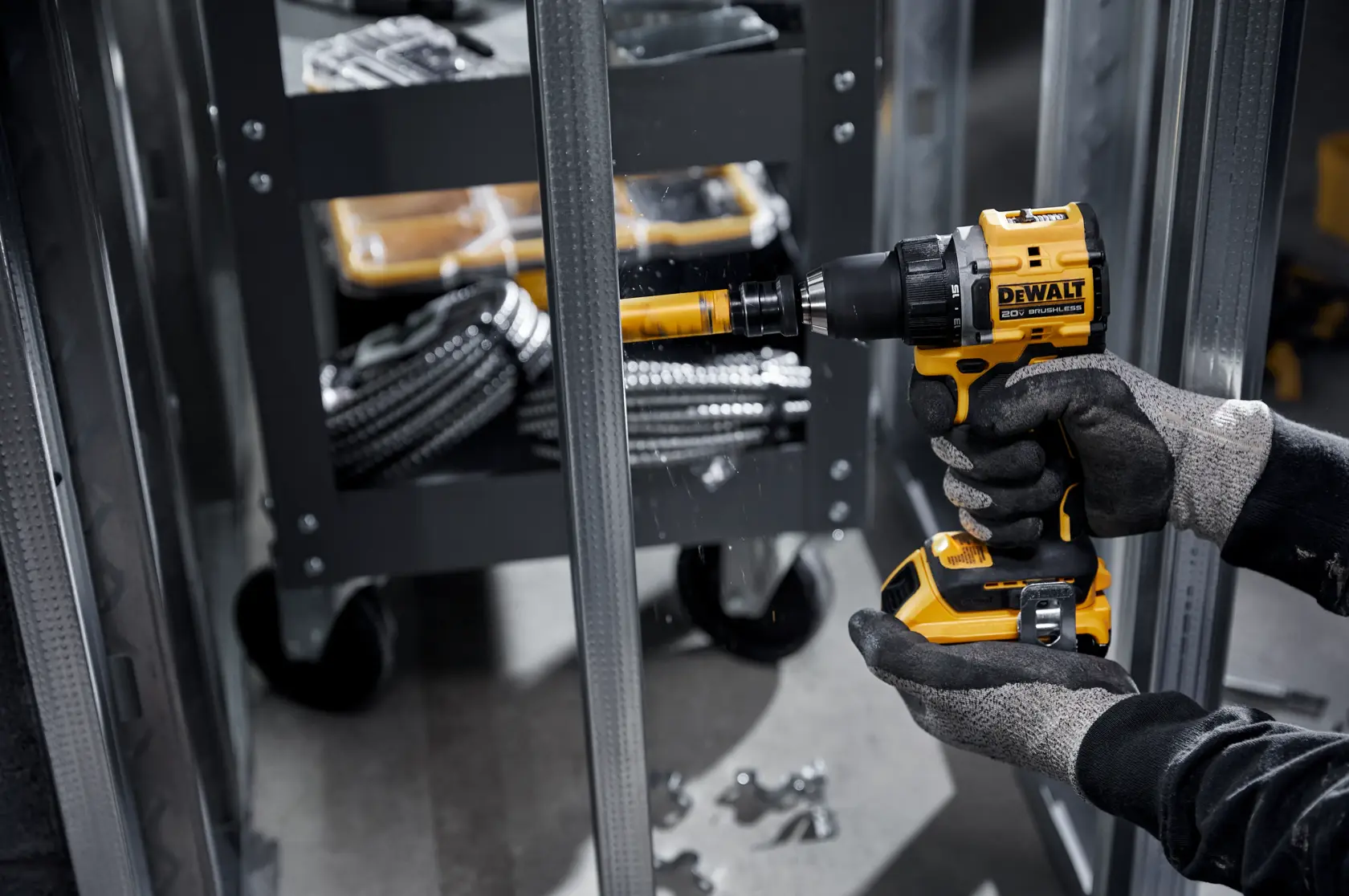A person wearing gloves is using a yellow and black DEWALT cordless drill to create a hole in a metal frame. In the background, there is a tool cart with various tools and coiled cables.