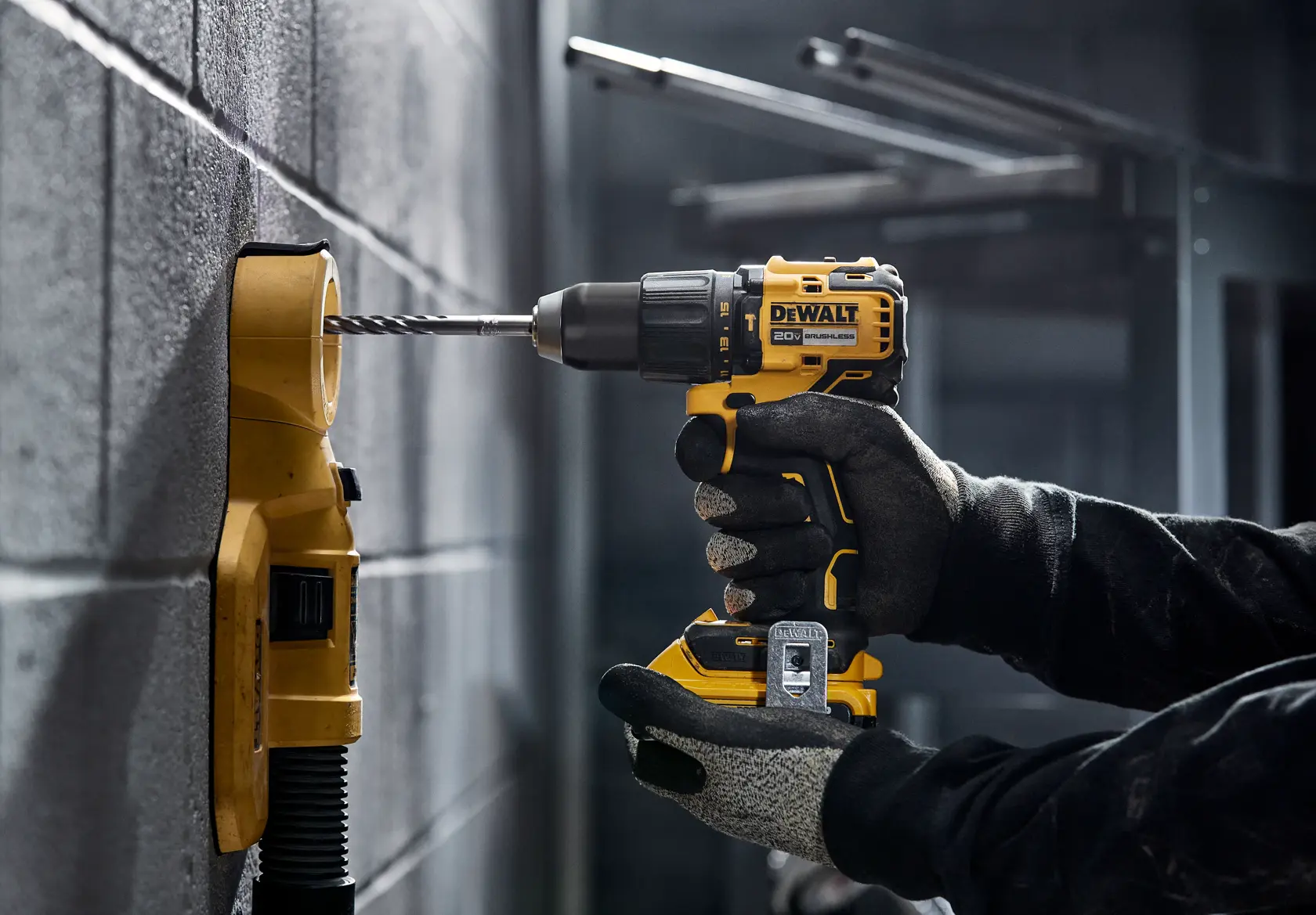 A person wearing gloves is using a yellow and black DEWALT cordless drill to drill into a concrete wall, with a dust collection attachment connected to the wall.