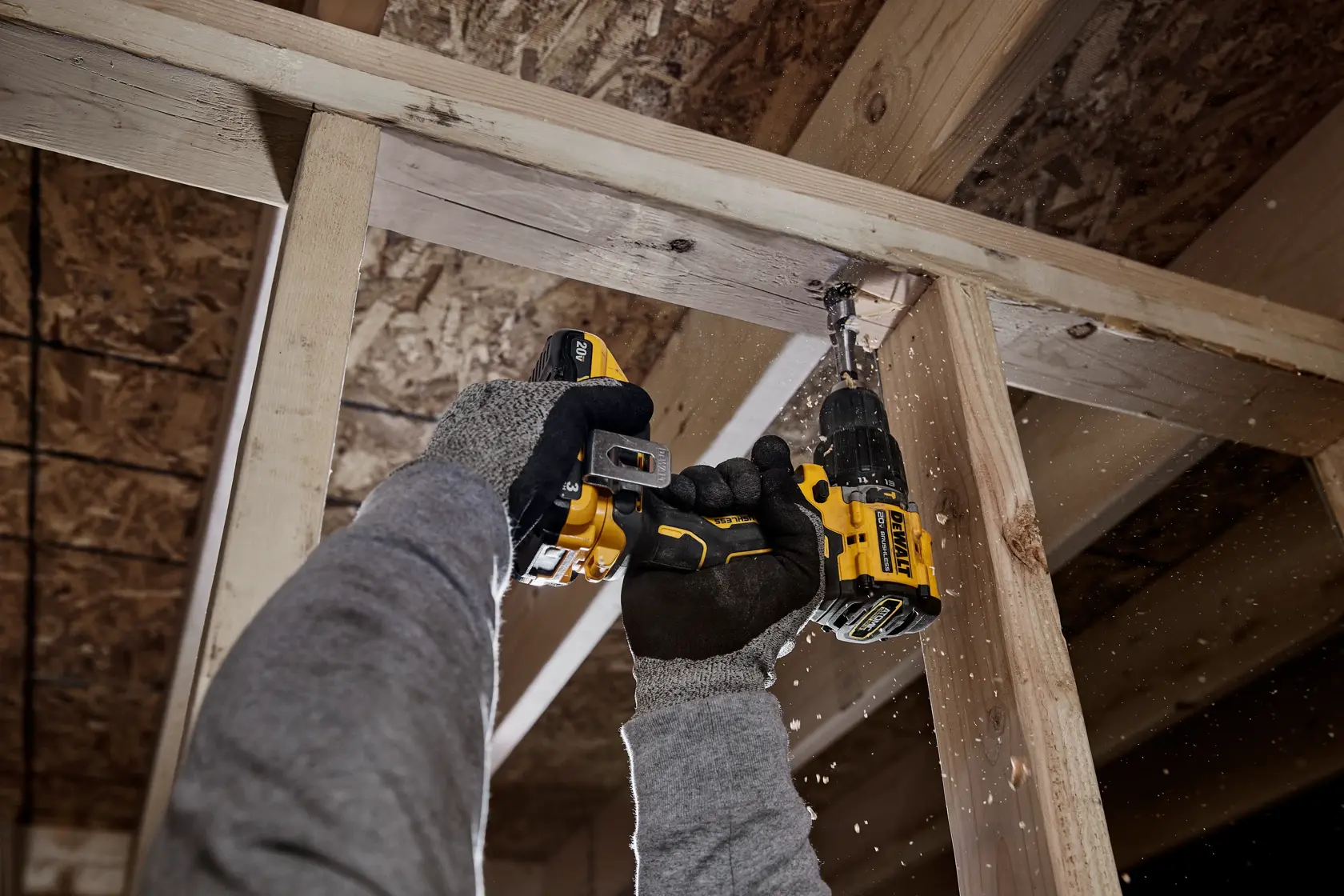 A person wearing gloves and a gray sweatshirt is using a DEWALT cordless drill to drill into a wooden beam inside a construction framework. Wood shavings are visible as the drill makes contact with the beam.