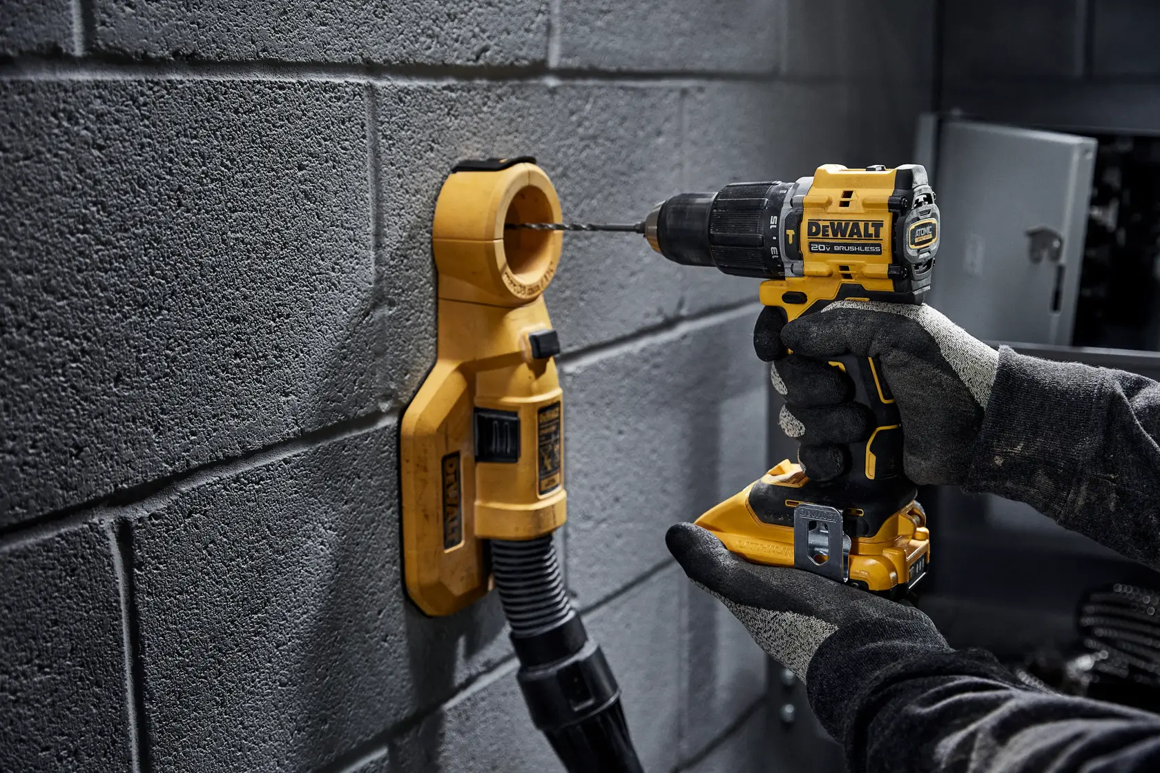 A person wearing work gloves is using a DEWALT cordless drill to drill into a concrete block wall, with a DEWALT dust collection attachment positioned around the drill bit to capture debris.