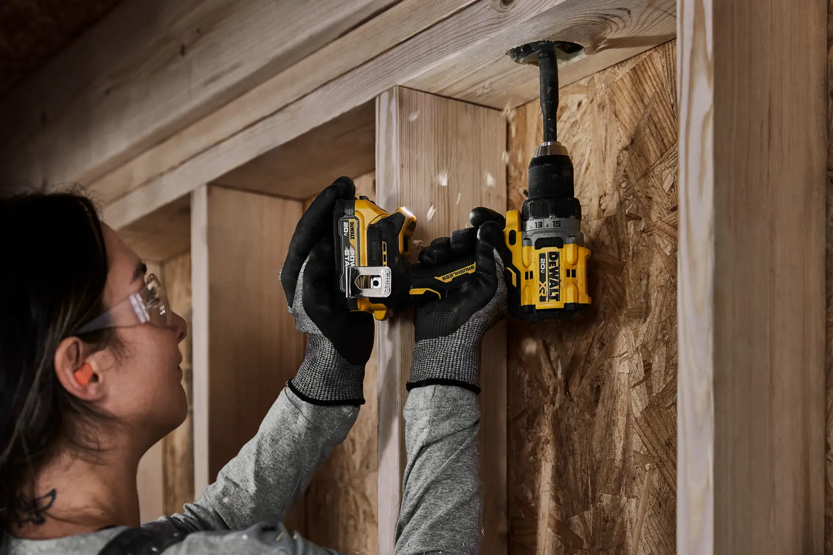 A person wearing work gloves uses a yellow cordless drill to bore a hole in a wooden wall frame. The person's face is blurred for privacy.