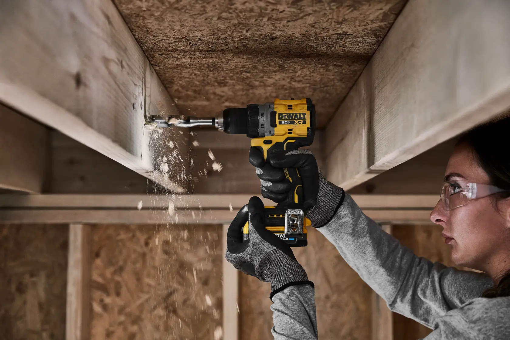 Person wearing gloves is using a yellow DeWalt cordless drill to drill into a wooden beam in a construction setting, with wood shavings falling.