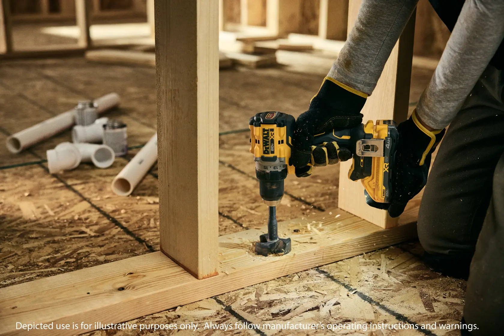 A person using a DEWALT cordless drill to bore a hole in a wooden beam during construction, wearing work gloves and kneeling on a plywood floor. Various PVC pipes and fittings are scattered in the background.