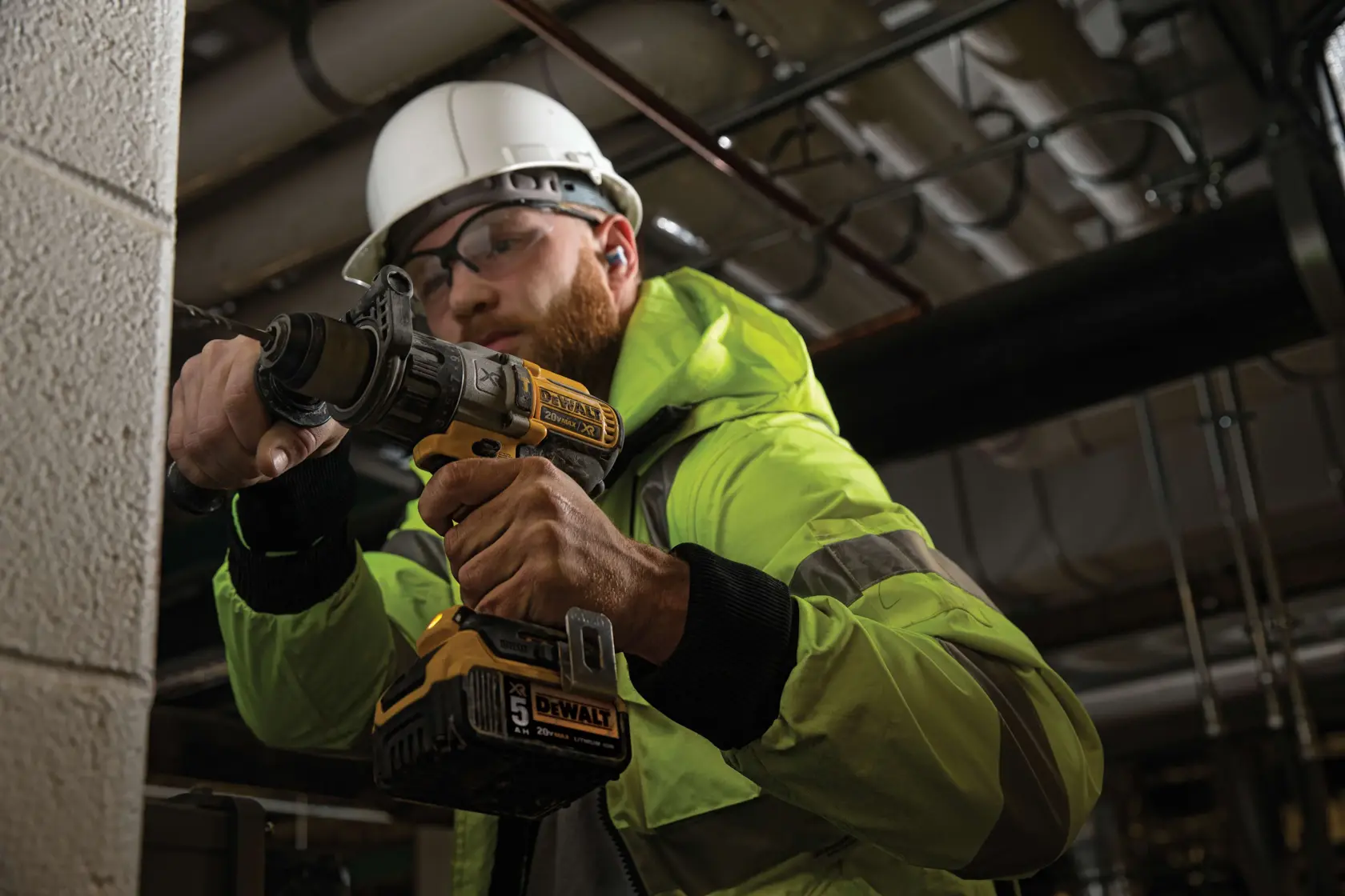 A person wearing a green safety jacket and white hard hat is using a DEWALT DCD996P2 cordless power drill to work on a concrete wall in an industrial setting. The face is obscured for privacy.