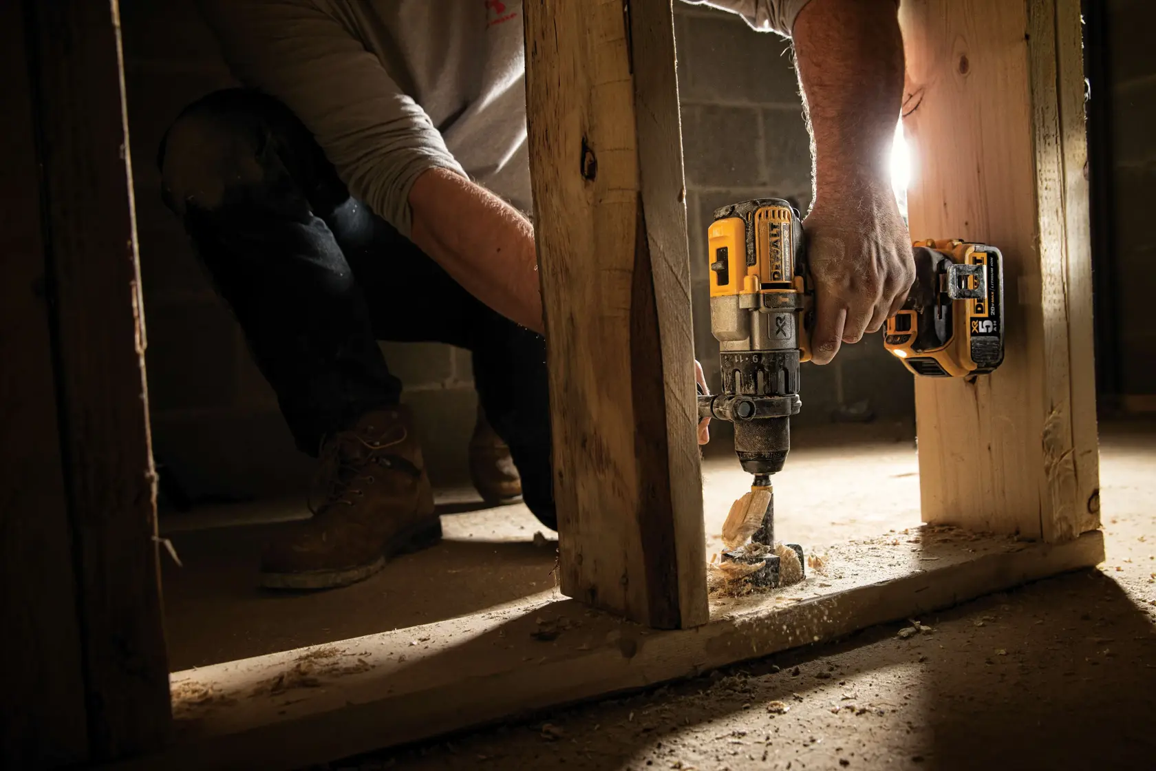 A person using a DEWALT DCD996P2 power drill to drill through a wooden beam at a construction site. There is sawdust on the floor and the person is wearing work boots and jeans.