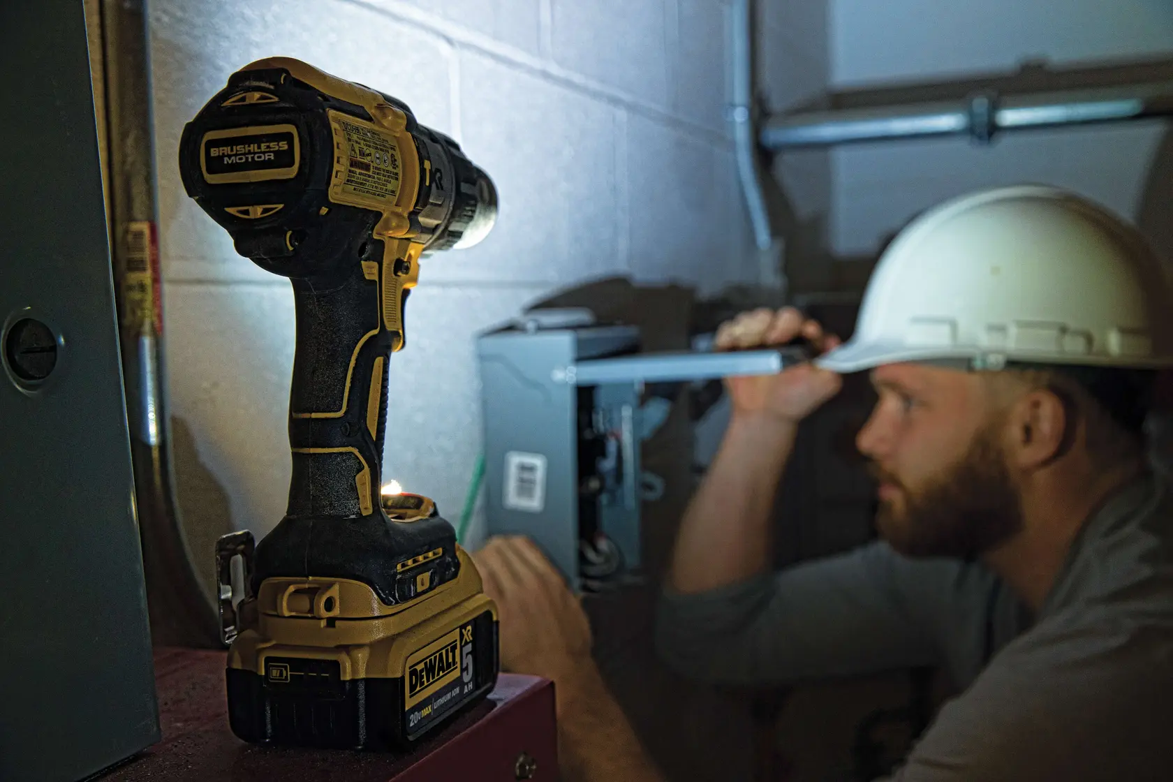 A DEWALT cordless drill with a brushless motor is shown in the foreground, placed on a surface, with a person wearing a hard hat working on electrical equipment in the background.