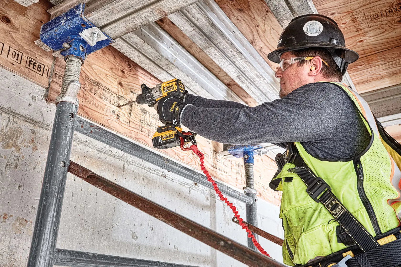 A worker wearing a safety vest and helmet uses a DEWALT DCD996P2 cordless drill to work on a wooden ceiling structure at a construction site. The worker is positioned on scaffolding.