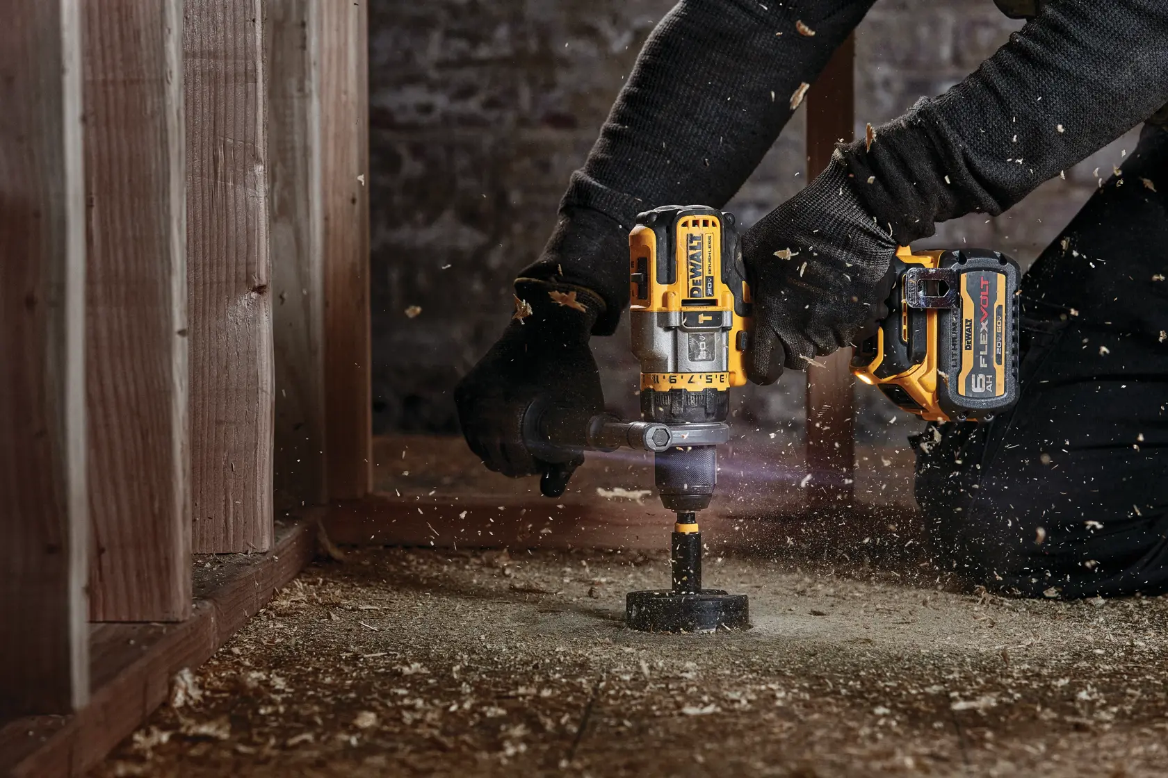 A person uses a DEWALT cordless power drill, model DCD999T1, to drill into a wooden floor. The person is wearing black gloves and working in a construction or renovation setting, with wood shavings flying around.