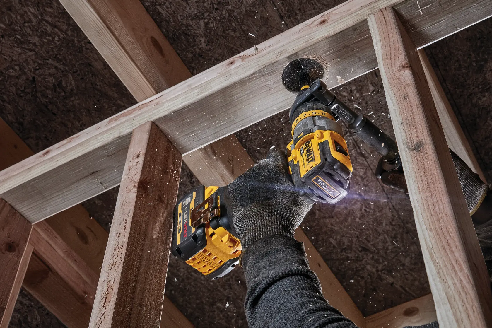 A person wearing work gloves uses a DEWALT cordless power drill to drill through a wooden beam as part of a construction project. Wood chips and dust are visible flying from the drilling area.