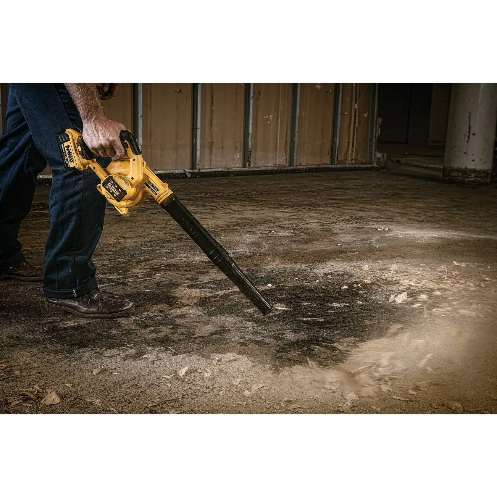 A person using a yellow DEWALT cordless blower to clear dust and debris from a concrete floor in an industrial or construction environment.