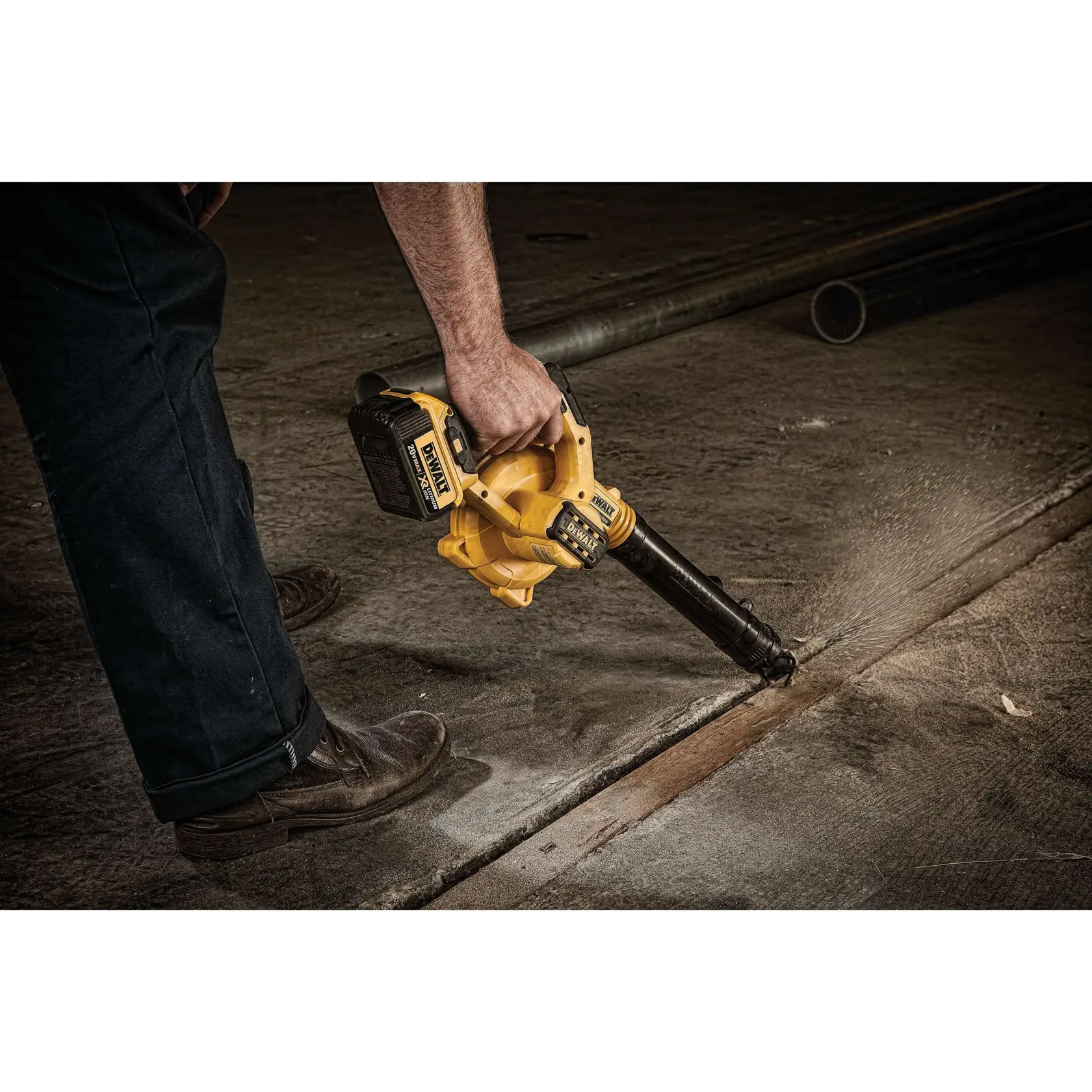 A person uses a DEWALT cordless handheld blower (SKU: DCE100M1) on a concrete floor to clear dust and debris.