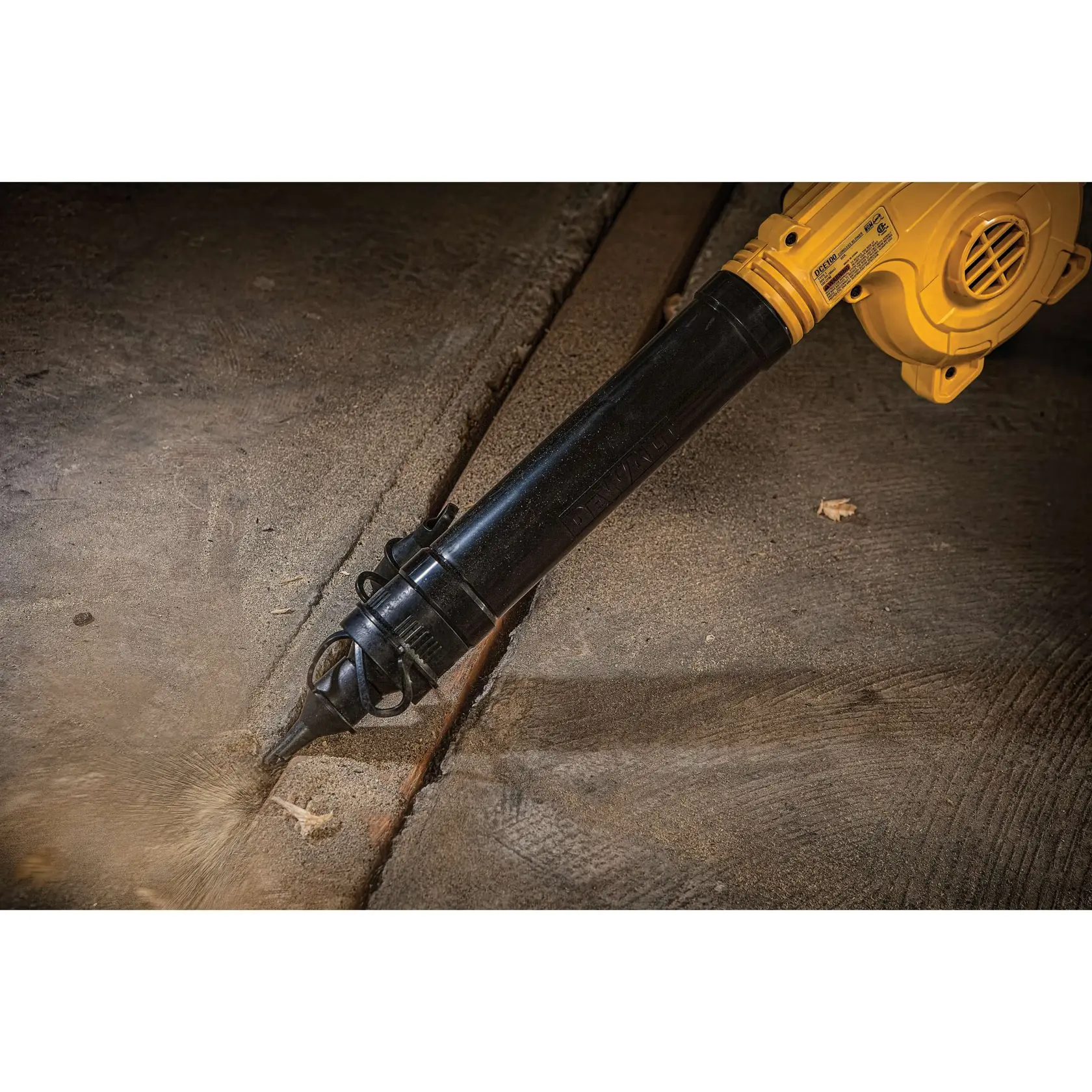 A yellow and black DEWALT cordless blower is being used to blow debris out of a crack in a concrete surface.
