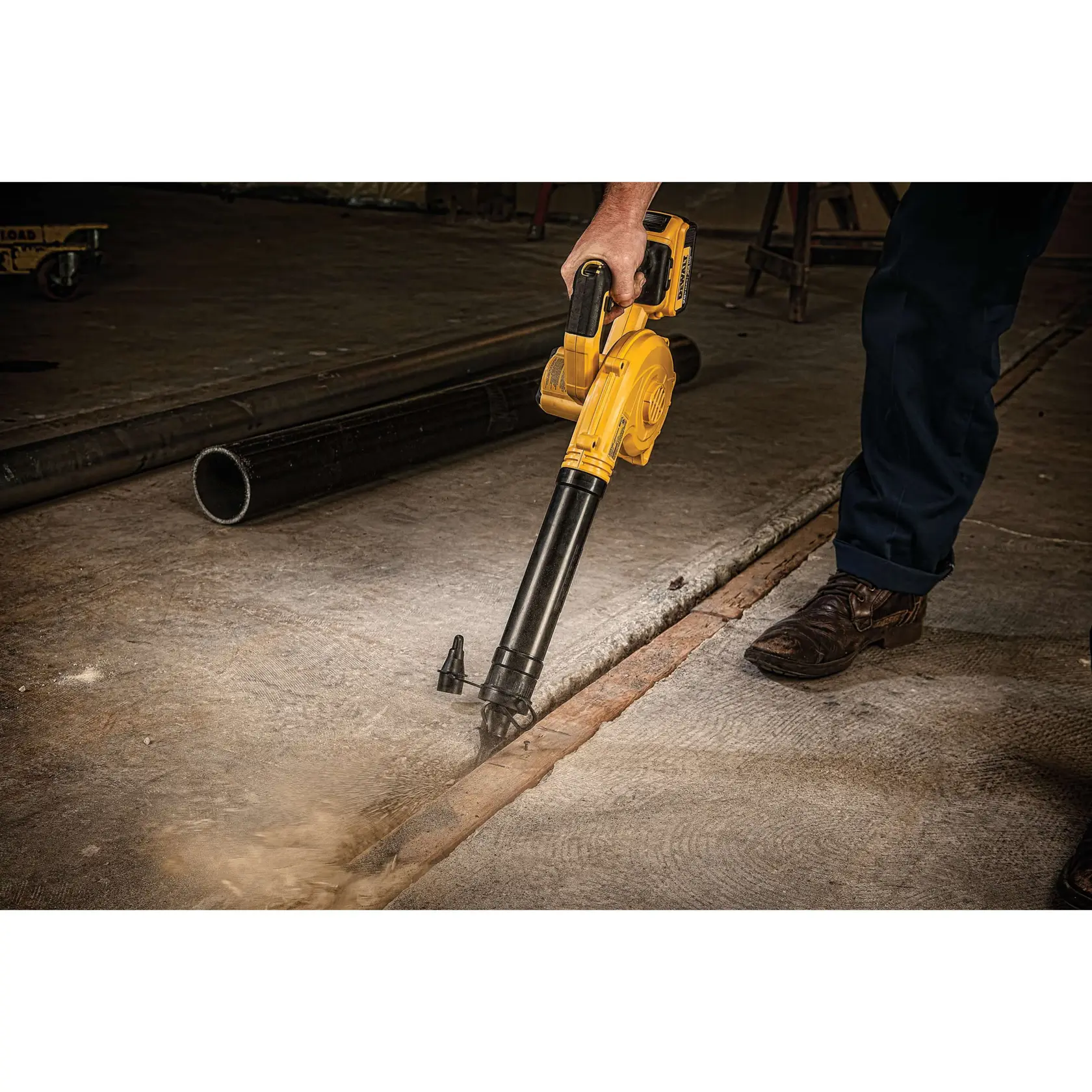 A person using a yellow DEWALT cordless blower to clear dust and debris from a concrete floor in an industrial setting.