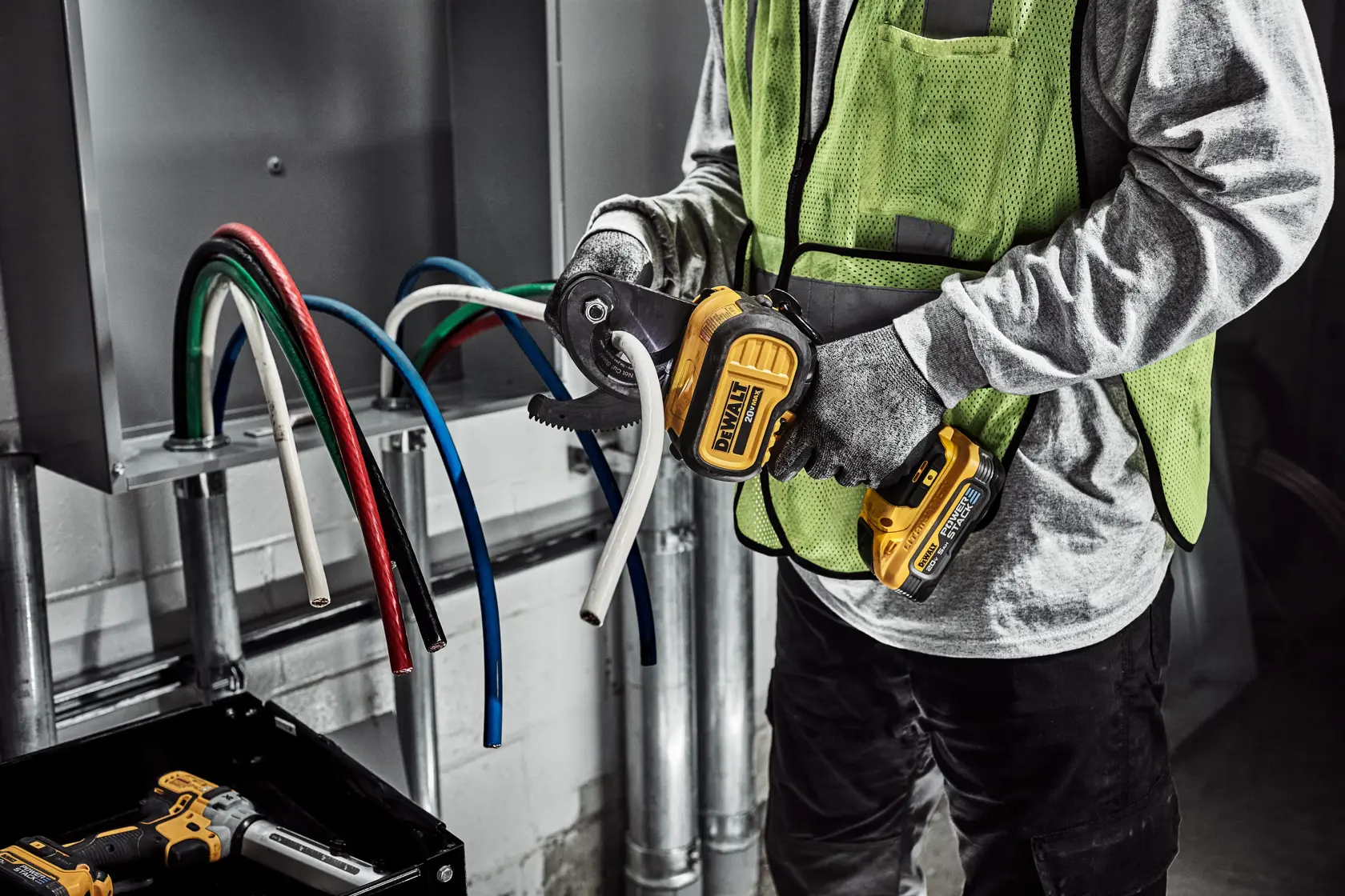 A person wearing gloves and a safety vest is using a DEWALT cordless cable cutter to cut a thick white electrical cable. Several colored cables are visible coming out of an electrical panel.