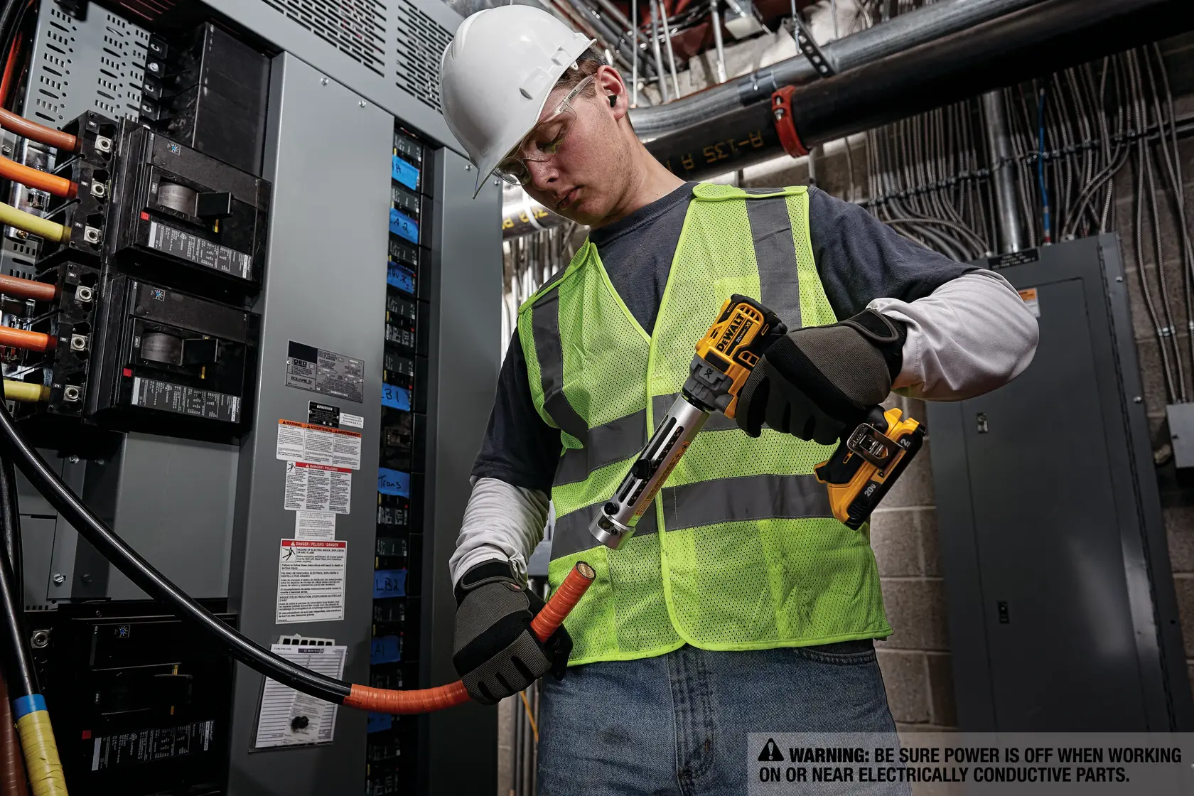 A worker wearing a white hard hat, safety gloves, and a neon green safety vest uses a DEWALT cordless tool to cut an electrical cable in an industrial setting. The worker is standing next to an electrical panel with multiple wires and breakers. A warning label is visible, advising to ensure power is off when working near electrically conductive parts.