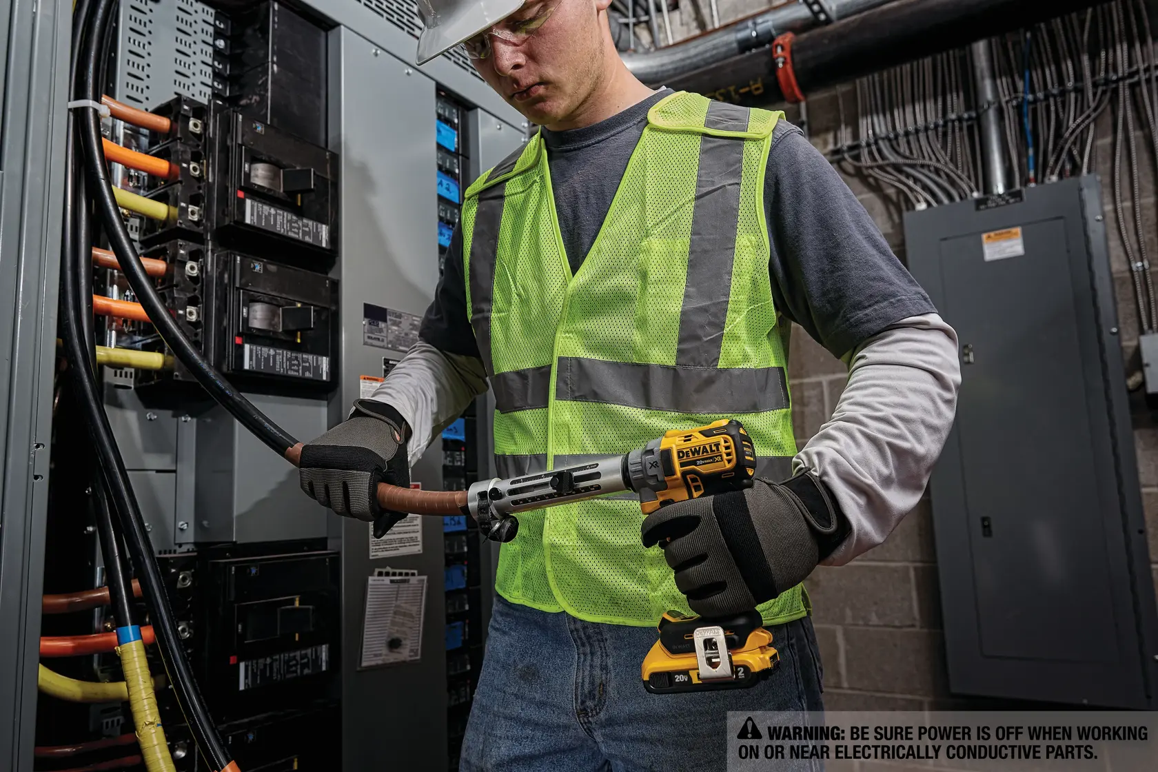 A person wearing a reflective safety vest and gloves is using a DEWALT cordless tool to work on thick electrical cables inside an electrical panel or breaker box. The environment appears to be an industrial or commercial electrical installation with warning signage visible.
