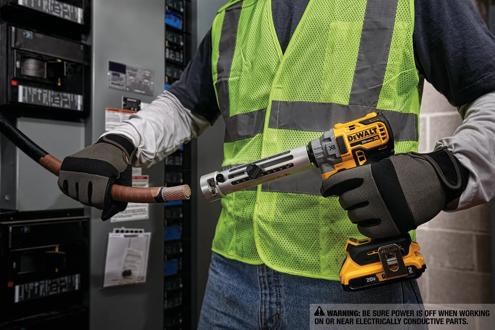 A person wearing safety gloves and a reflective vest uses a DEWALT power tool to strip insulation from a thick electrical cable in front of an electrical panel.