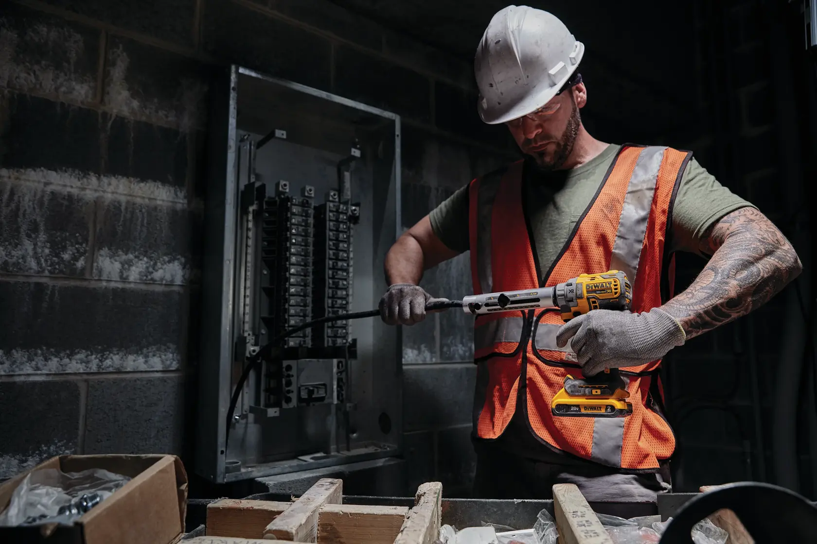 A worker wearing a hard hat and orange safety vest uses a DEWALT cordless tool to work on electrical wiring near a circuit breaker panel in an industrial setting.