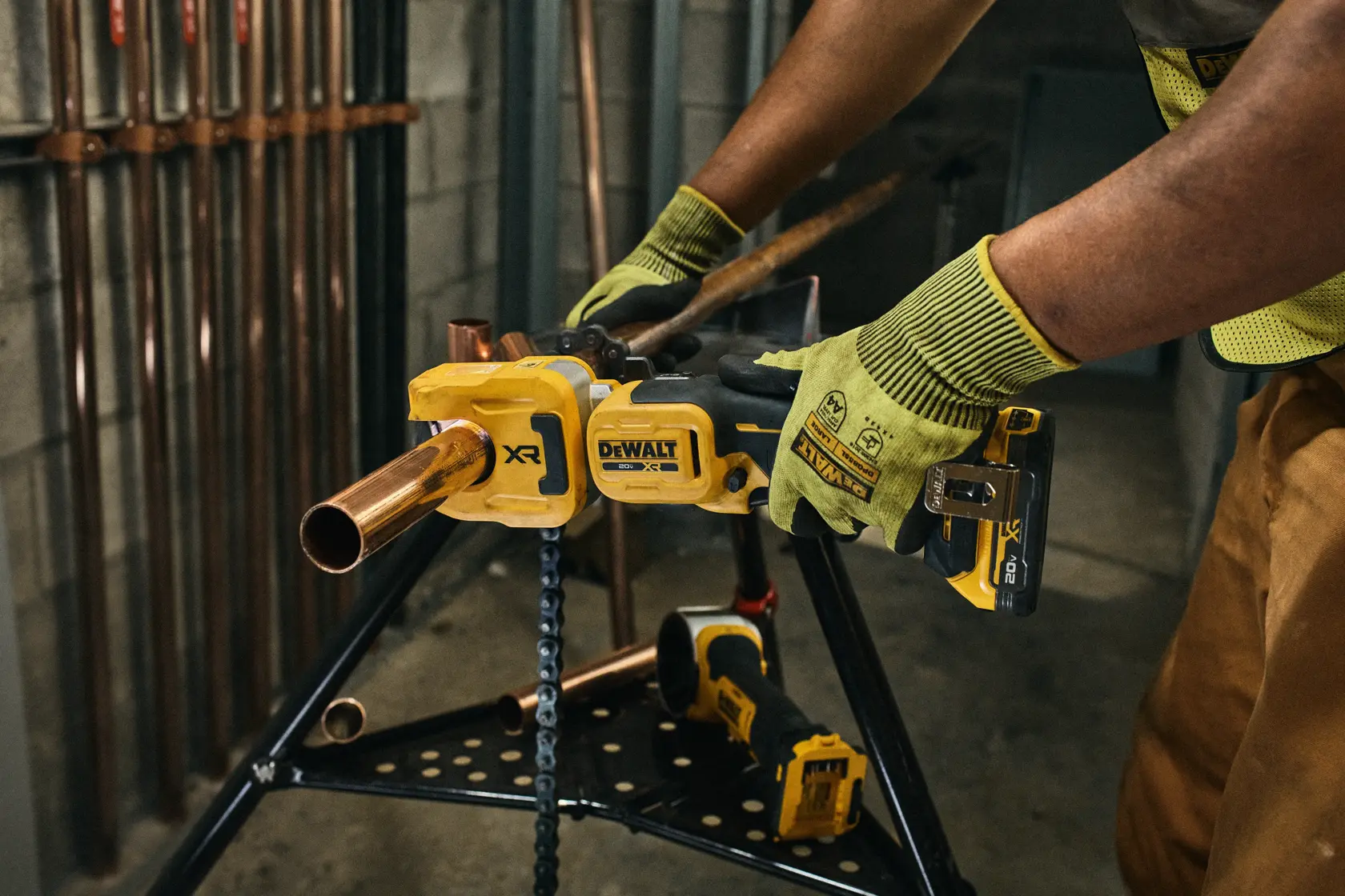 A person wearing gloves and a cap is using a DEWALT cordless pipe cutter to cut a metal pipe in an industrial workshop setting. The tool is yellow and black and features the DEWALT branding.