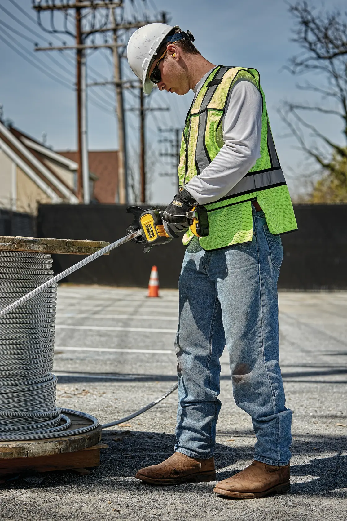 A person wearing a hard hat, high-visibility vest, gloves, and jeans is using a DEWALT power tool to work with a large spool of cable outdoors in a parking lot.