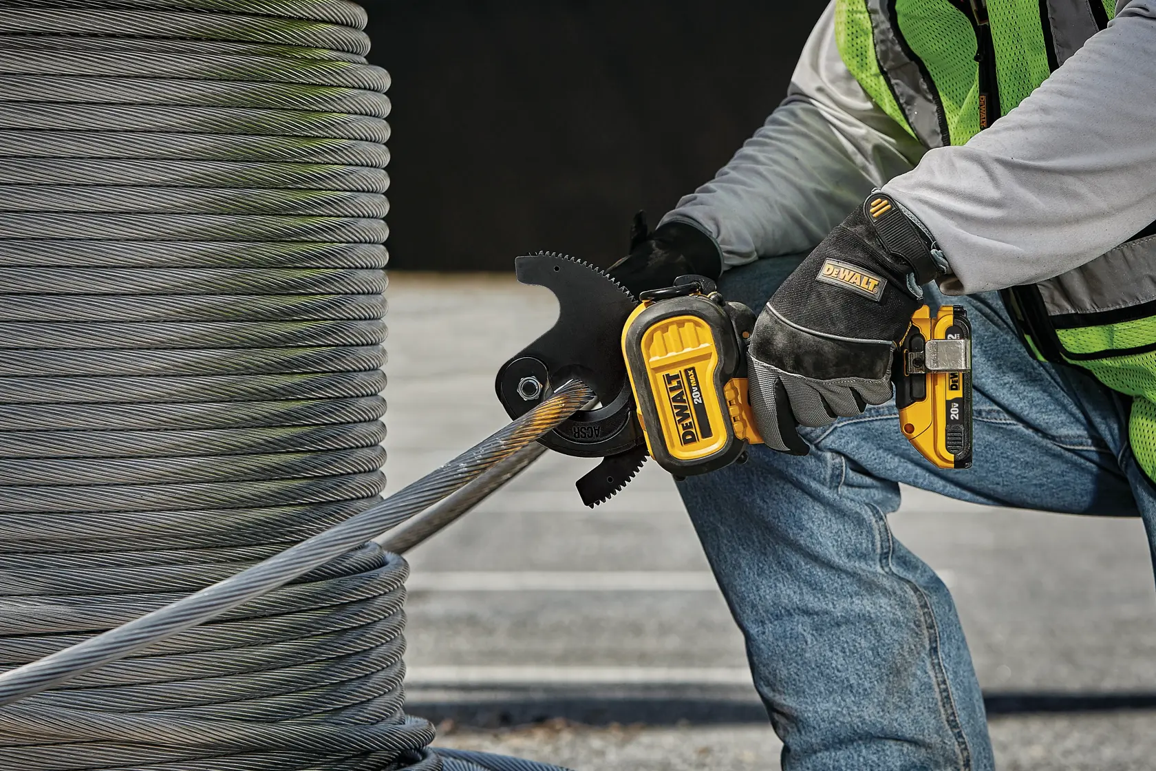 A person wearing safety gloves and a safety vest uses a DEWALT cordless power tool to cut through a thick metal cable next to a large spool of wire outdoors.