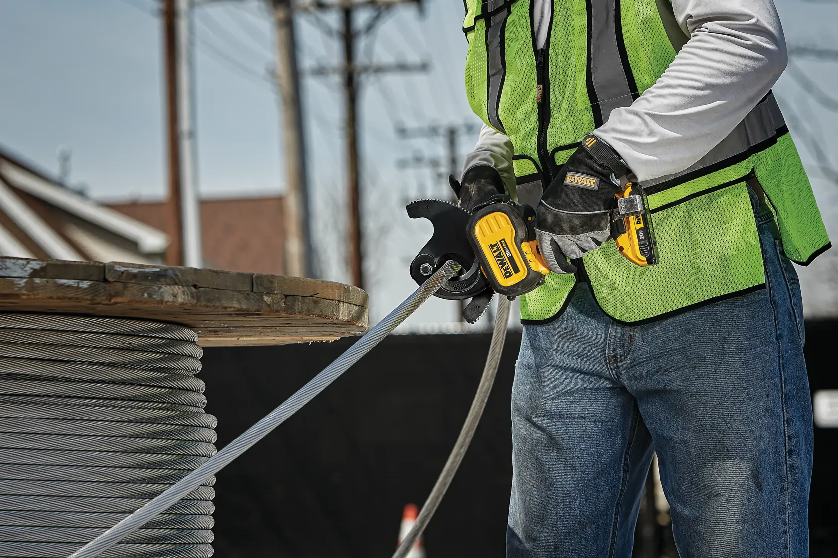 A person wearing a safety vest and gloves is using a DEWALT cordless cable cutter to cut a thick metal cable from a spool, outdoors at a work site.