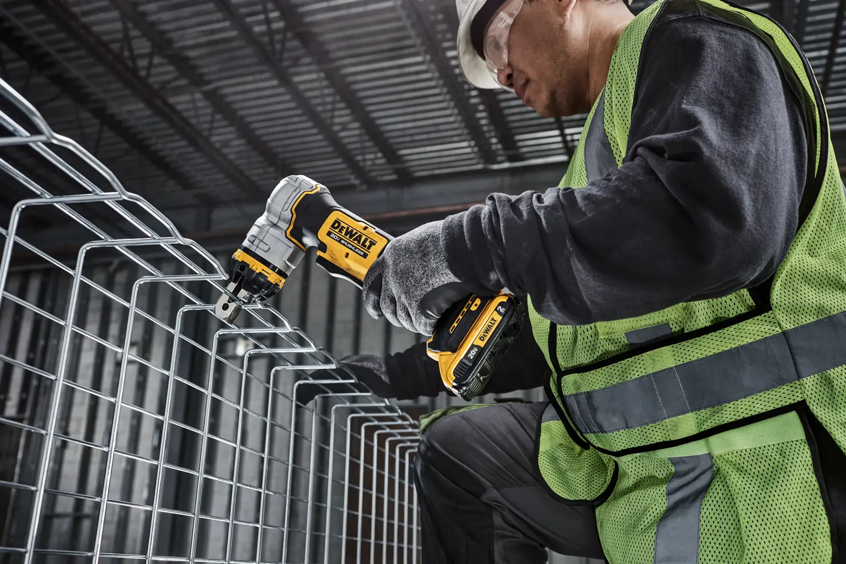 A person wearing a safety vest and gloves uses a DEWALT cordless metal shear to cut wire mesh in an industrial setting. The face of the person is blurred for privacy.