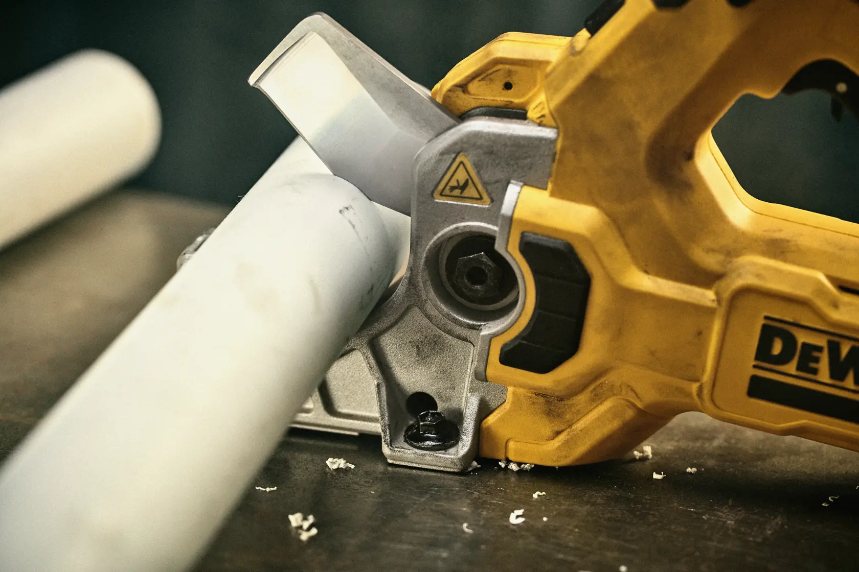 Close-up of a DEWALT handheld power tool cutting a white plastic pipe on a workbench. The tool is yellow and silver, with plastic shavings visible on the surface.