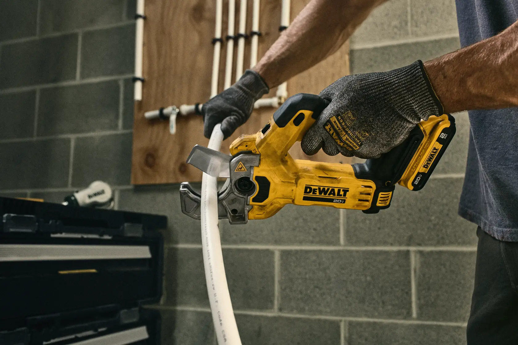 A person wearing work gloves is using a yellow DEWALT cordless pipe cutter to cut a white plastic pipe in an industrial setting with a concrete block wall and toolboxes.