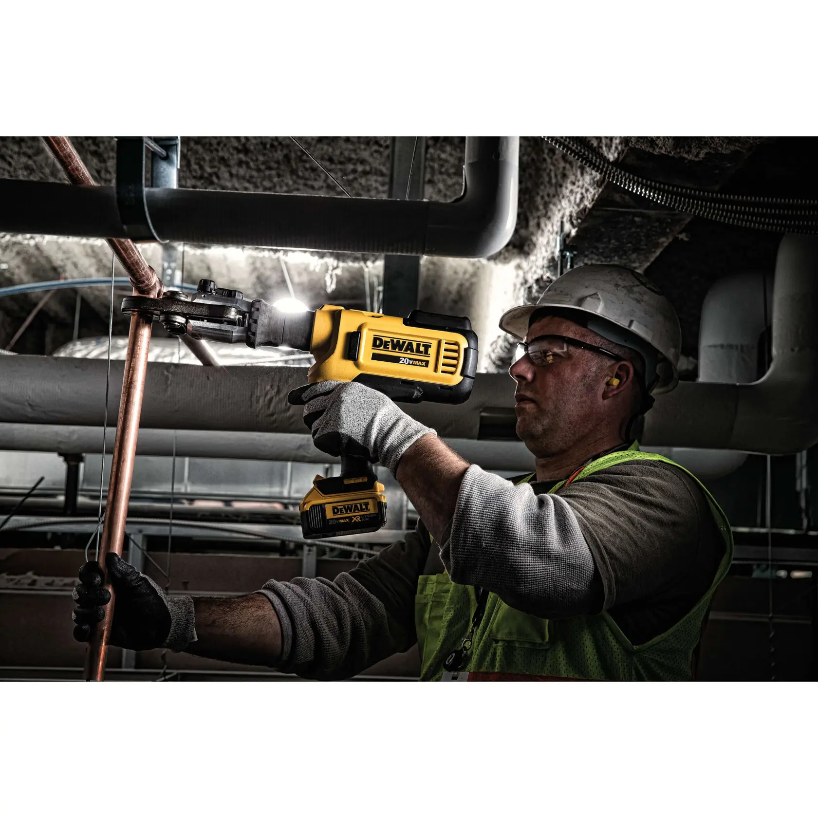 A worker wearing gloves, a high-visibility vest, and a hard hat is using a DEWALT DCE200M2K cordless tool to crimp a copper pipe in an industrial setting. The tool is yellow and black and features DEWALT branding.