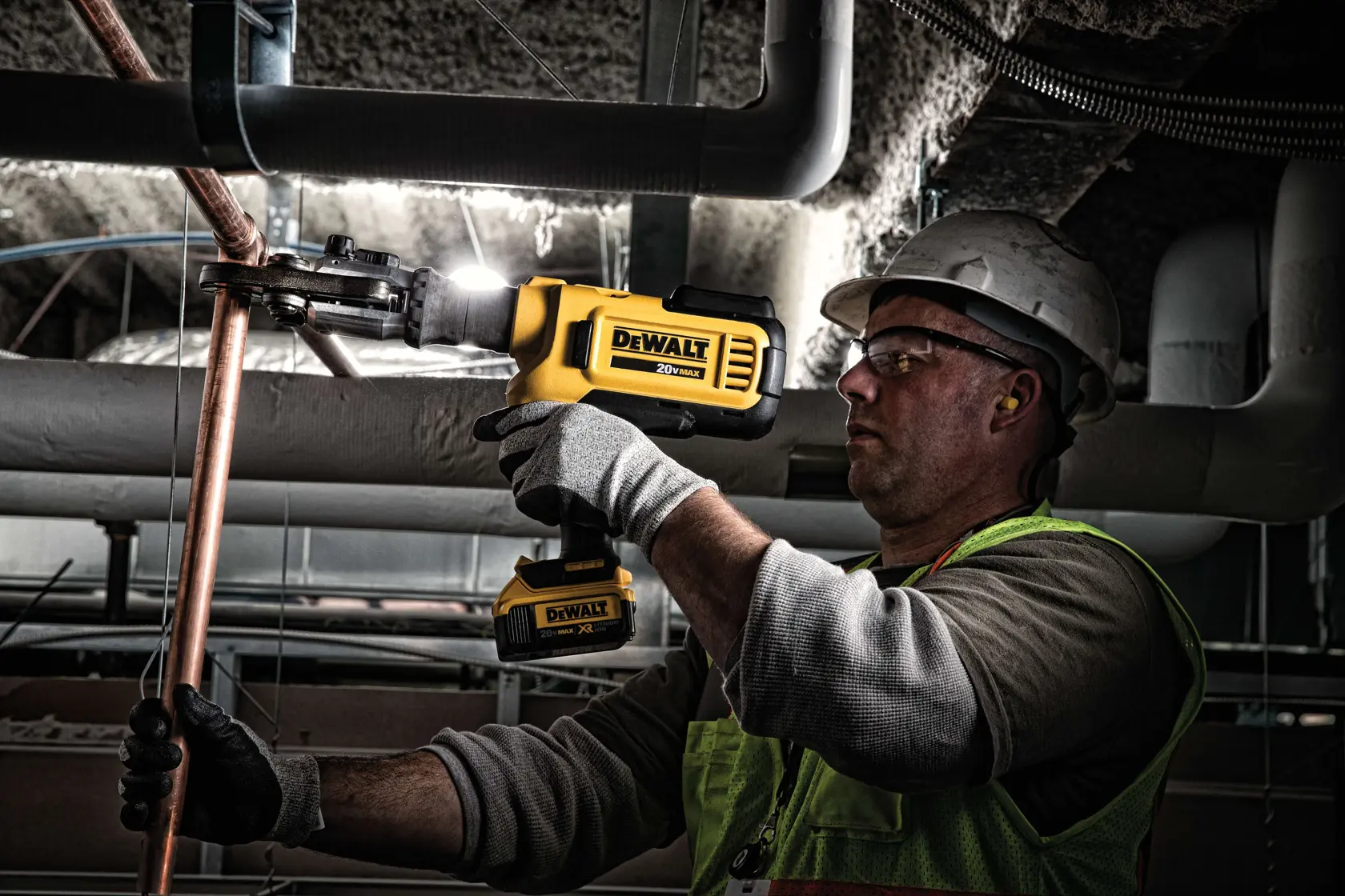 A person wearing safety gear uses a yellow DEWALT cordless tool to cut or shape a copper pipe in an industrial setting.