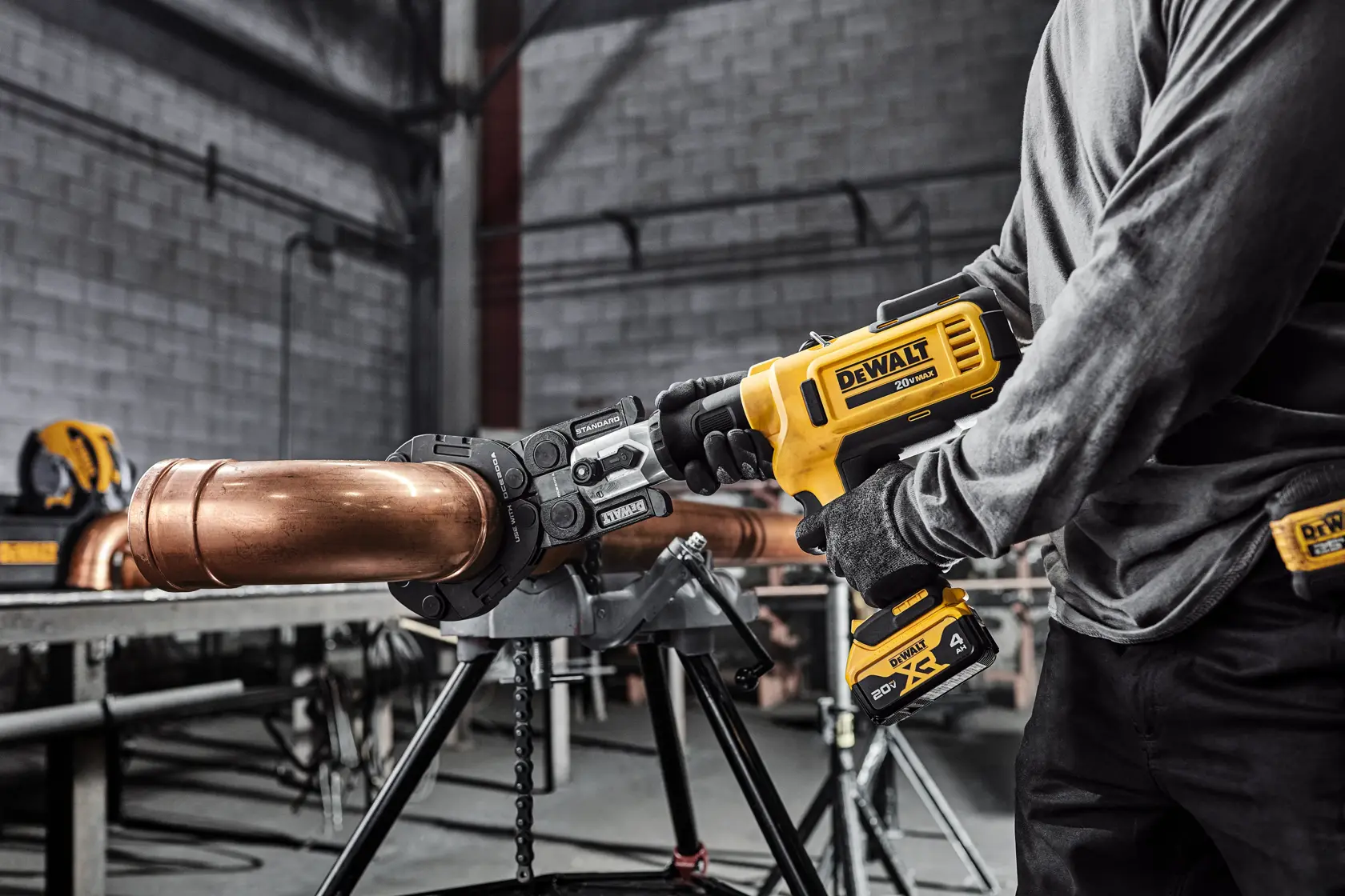 A person uses a DEWALT cordless tool to work on a large copper pipe secured in a metal stand inside an industrial workshop.