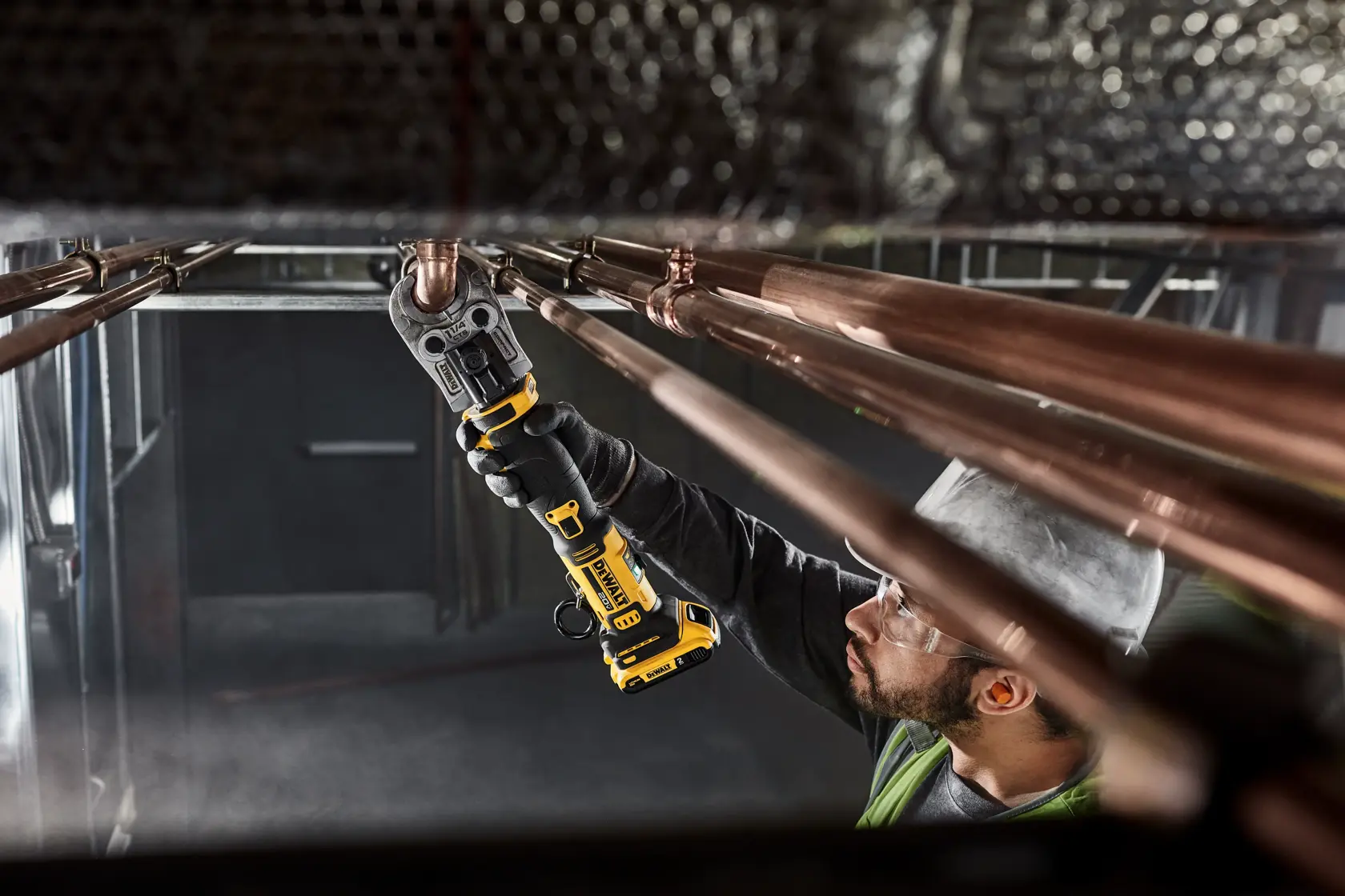 A person wearing a hard hat is using a DEWALT cordless tool (SKU: DCE210D2) to work on copper pipes in an industrial setting. The tool is yellow and black, and appears to be used for pressing or joining pipe fittings.