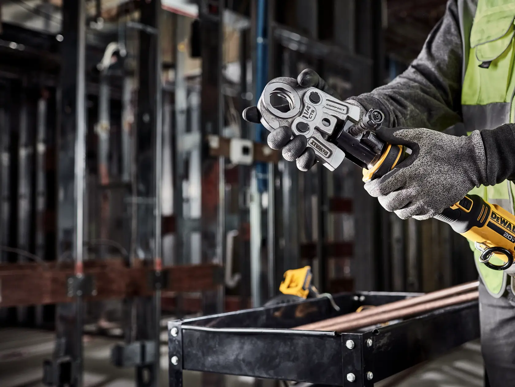Person wearing gloves operates a DEWALT cordless power tool, specifically the DCE210D2 model, in an industrial setting with metal pipes and a workbench visible.