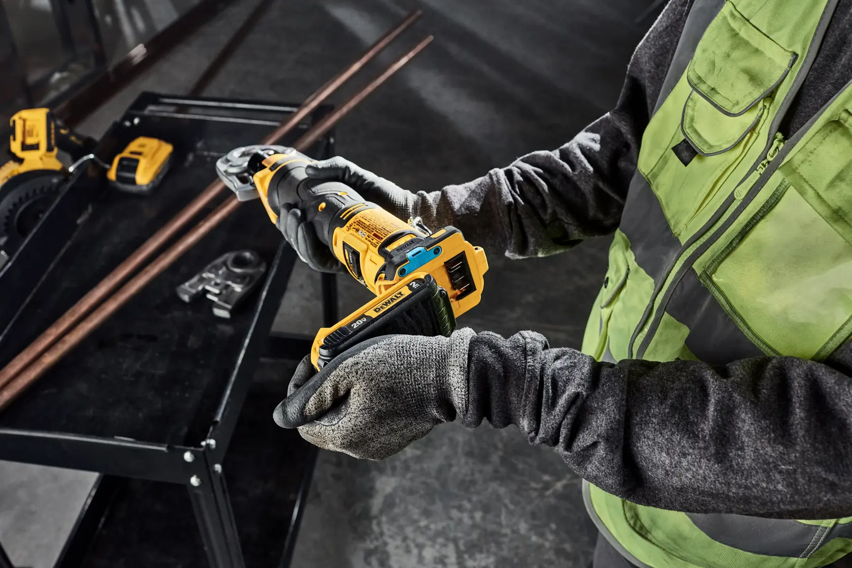 A person wearing a green vest and gloves is holding a DEWALT cordless tool, preparing for use. Nearby, copper pipes and other tools are placed on a black workbench in an industrial setting.