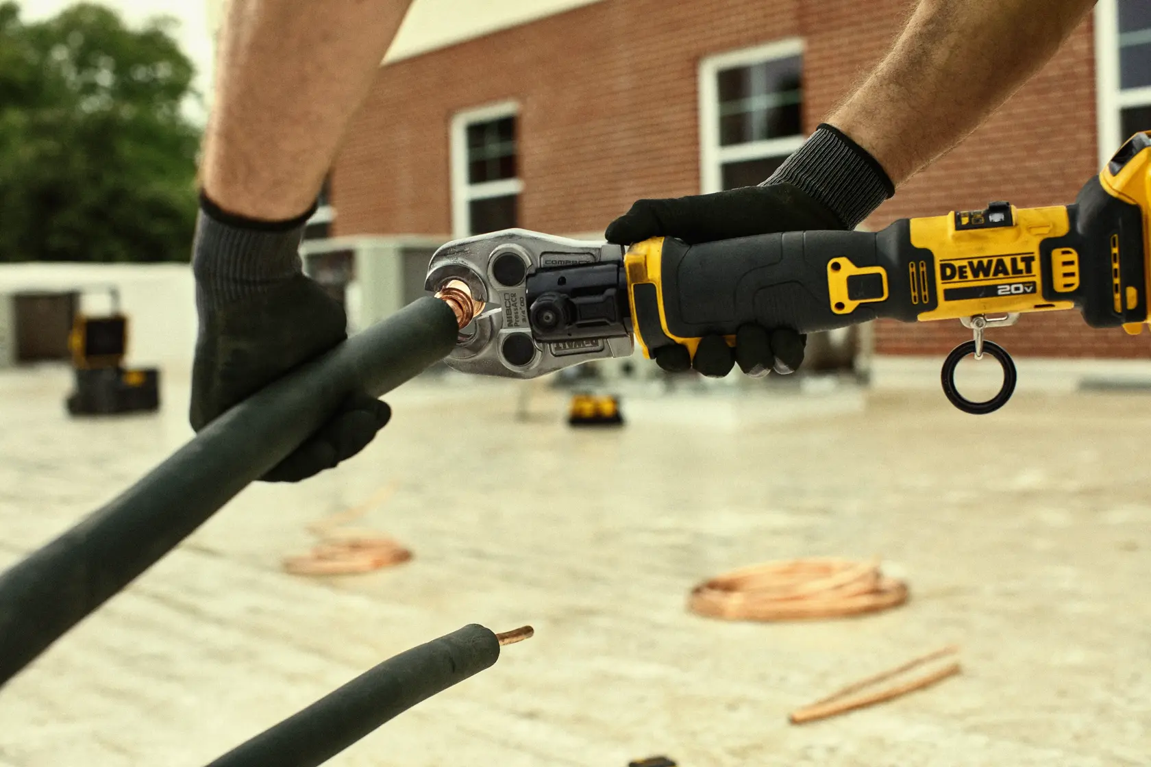 A person wearing gloves uses a DEWALT 20V cordless cutting tool to cut through a thick insulated cable outdoors, with a brick building and equipment visible in the background.
