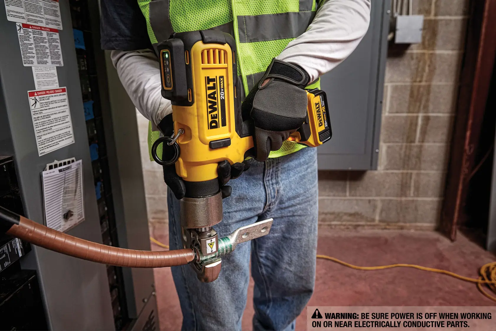 A person wearing a safety vest and gloves is using a yellow DEWALT DCE300M2 cordless tool to crimp a cable near an electrical panel. There is a caution warning about ensuring power is off when working on or near electrically conductive parts.