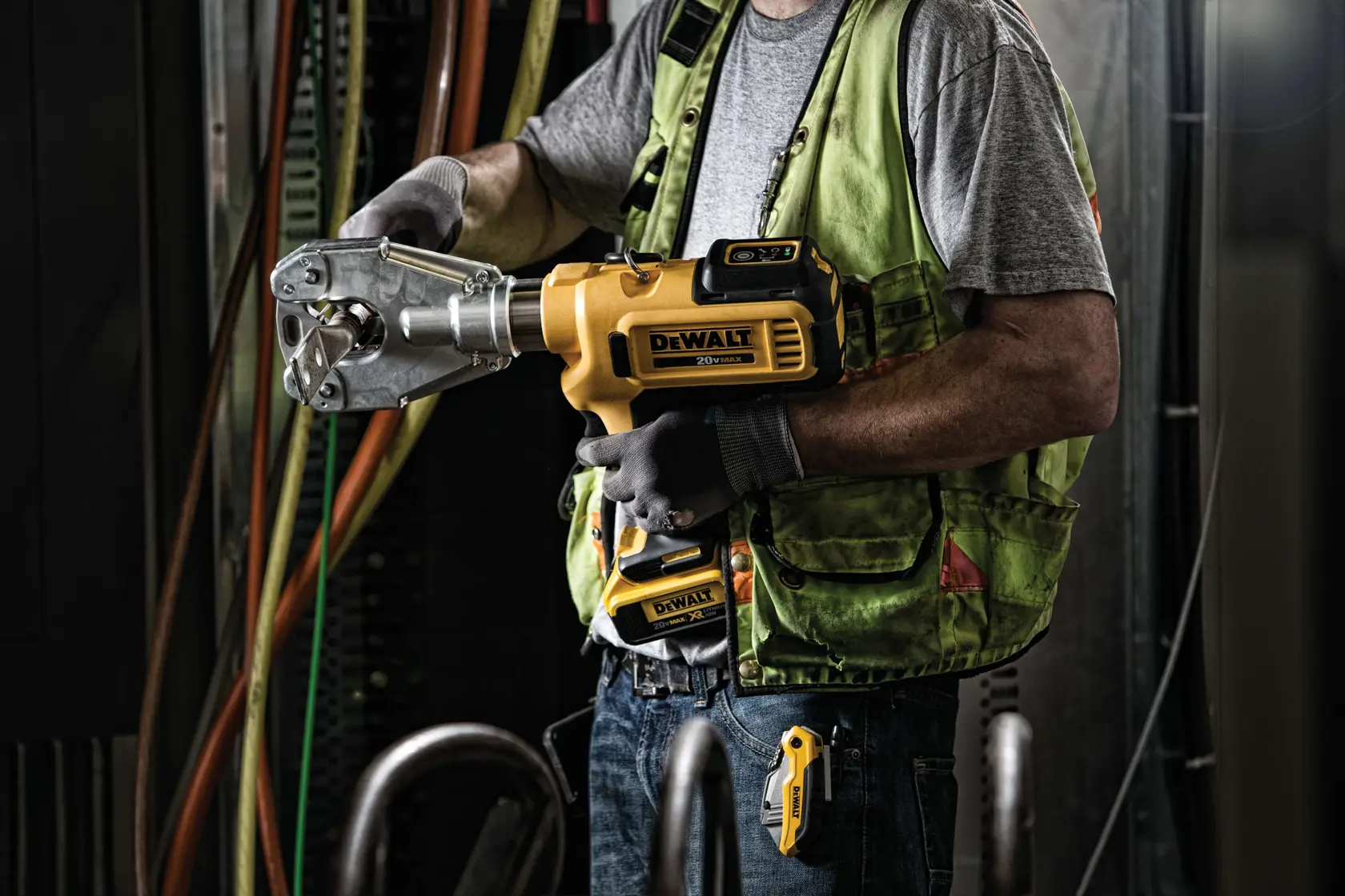 A person wearing a green safety vest and gloves is using a DEWALT battery-powered crimping tool, model DCE350M2, in an industrial setting with cables and equipment in the background.