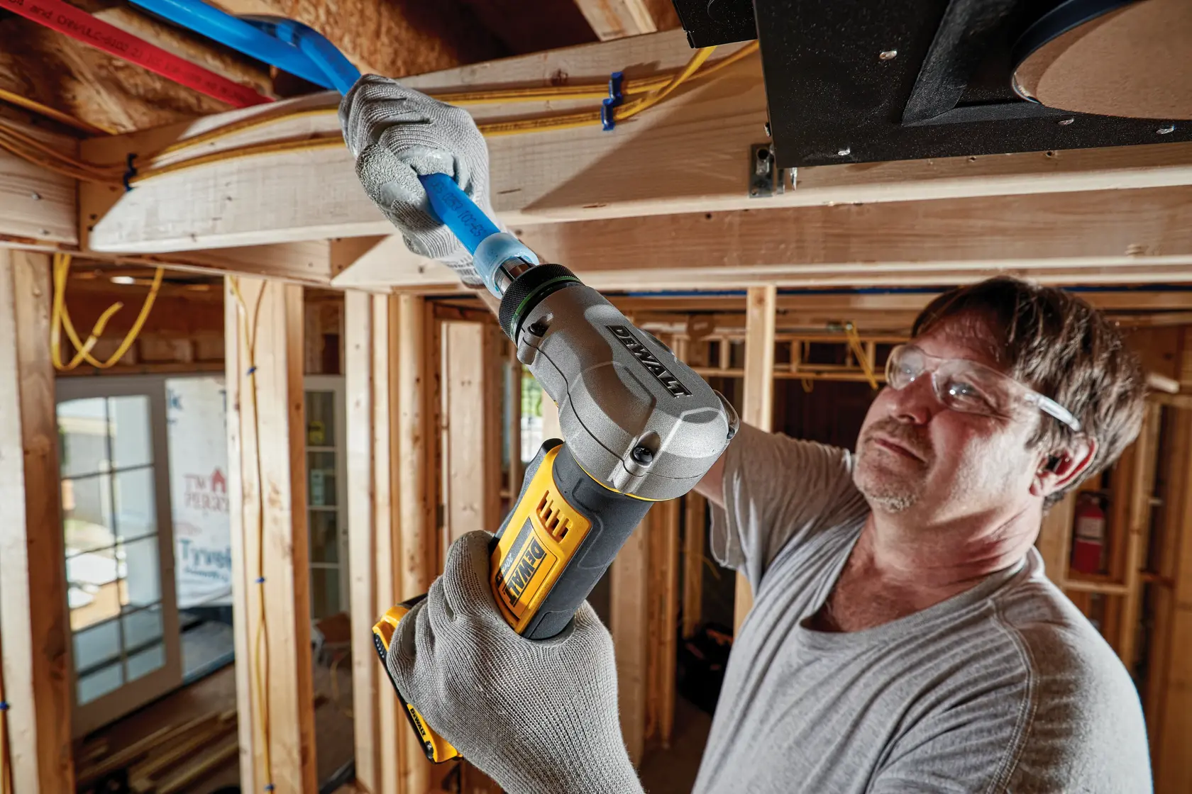 A person wearing gloves uses a DEWALT cordless tool to work on blue piping in a wooden construction site. The person's face is blurred for privacy.
