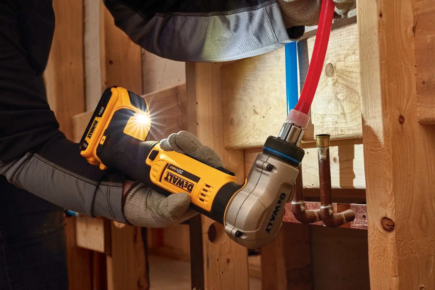 A person wearing gloves uses a yellow DEWALT cordless press tool to connect a pipe inside a wooden framework, with a bright LED light shining from the tool.