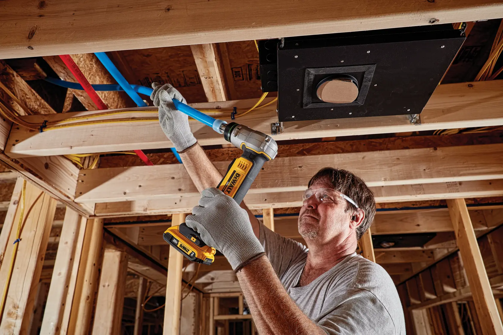 Person wearing gloves uses a DEWALT battery-powered tool to work on blue piping in a wooden-framed construction site.