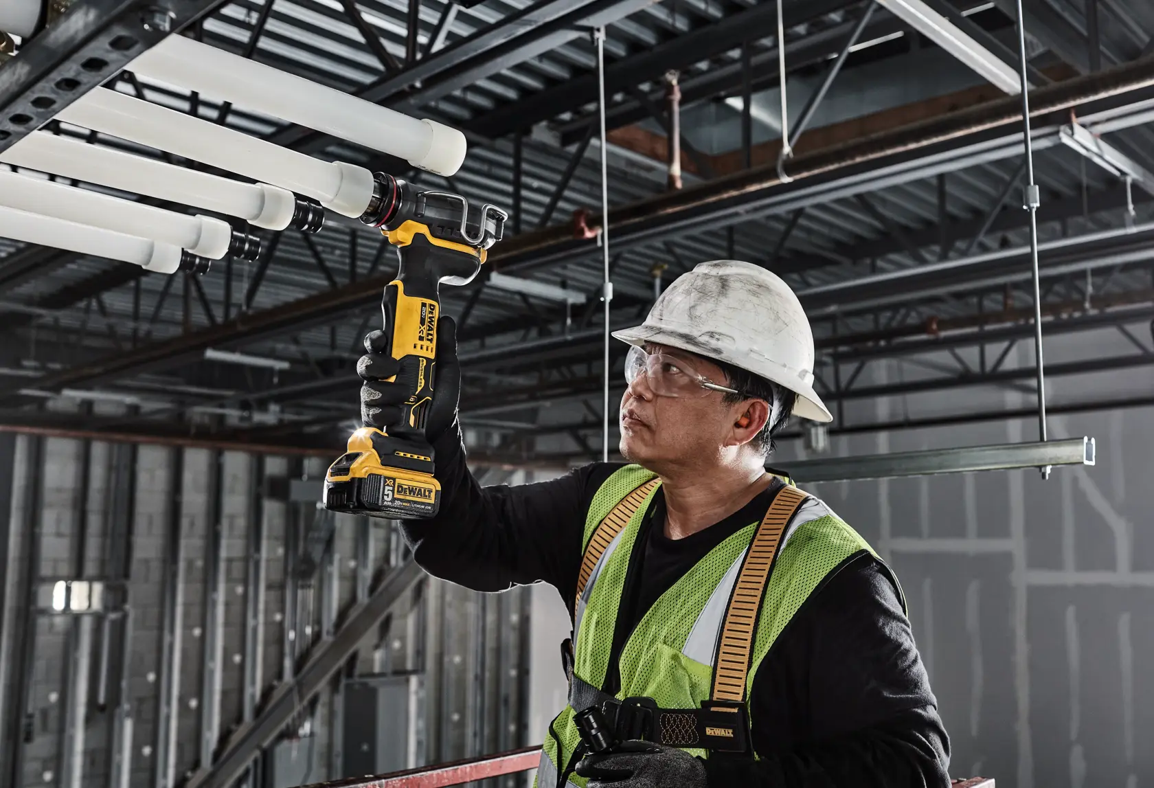 A construction worker wearing a safety vest and hard hat uses a DEWALT DCE410P1 inspection camera to examine pipes on a ceiling in an industrial building.