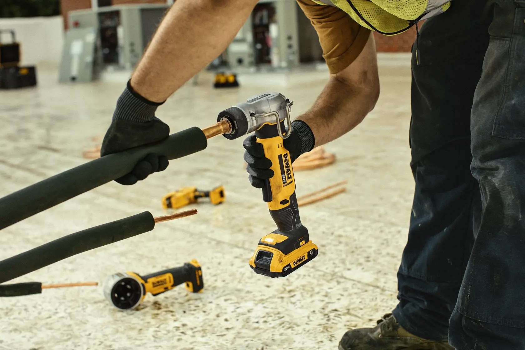 A worker uses a DEWALT cordless right angle drill to work on pipes in an industrial setting. The worker is wearing gloves and a yellow safety vest. Other DEWALT tools are visible on the floor.