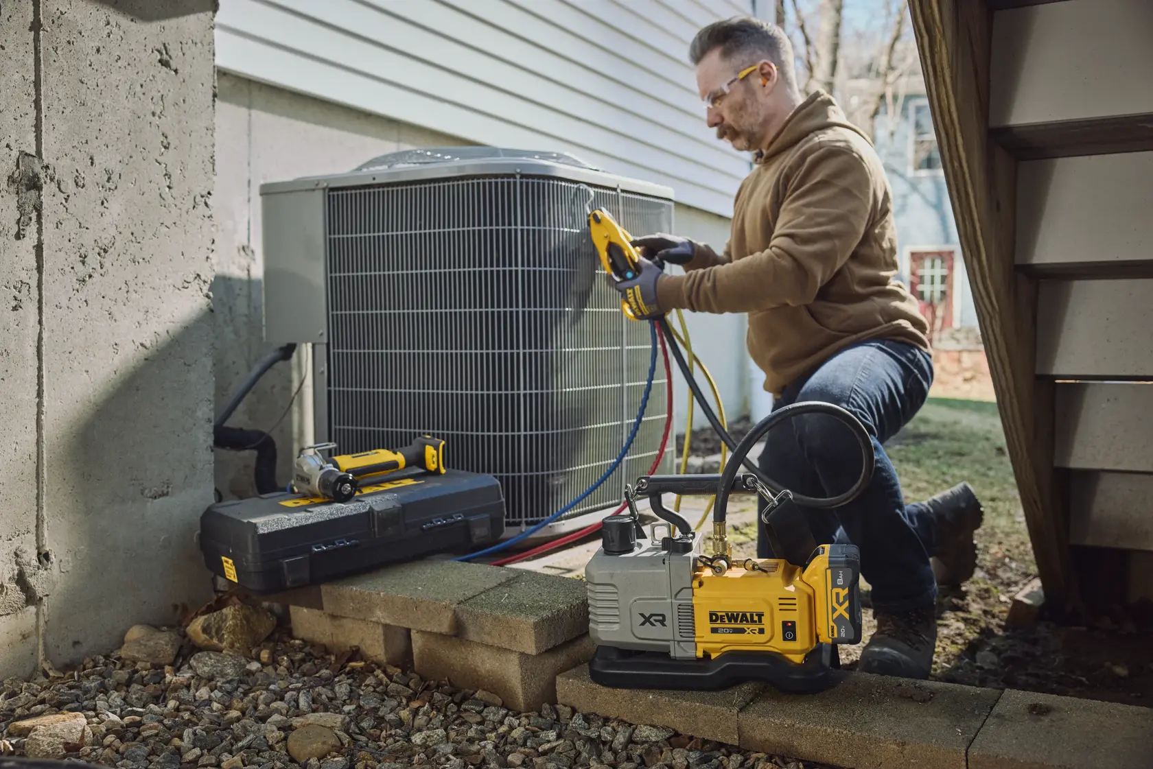 A person kneeling outside next to an air conditioning unit, using a DEWALT vacuum pump and tools for HVAC maintenance. DEWALT branded equipment and tool case are visible in the foreground.