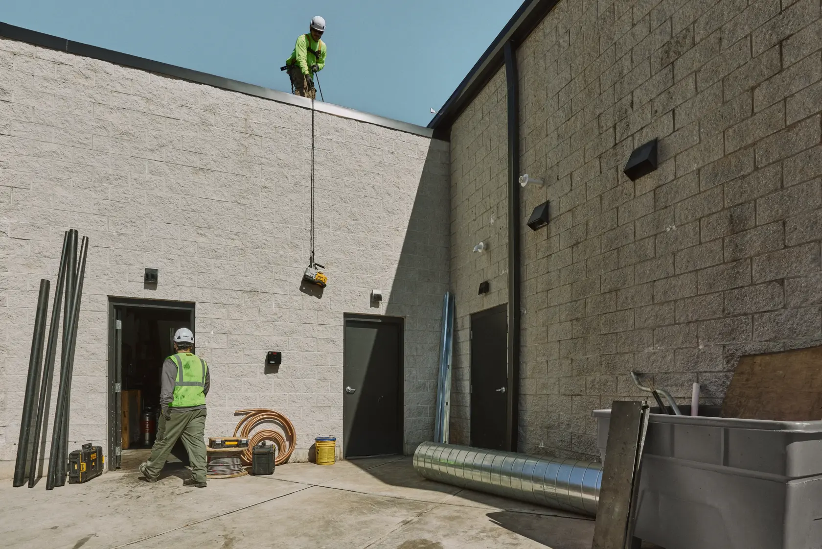 Two construction workers in safety gear are seen outside a building. One worker stands on the roof, lowering a DEWALT power tool with a rope, while the other worker waits below. Construction materials and equipment are scattered around the concrete yard.