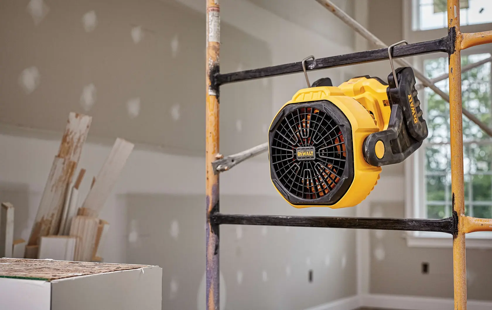 A yellow DEWALT portable fan, model DCE511C1, is attached to a metal scaffold inside a construction or renovation site. Unfinished drywall panels and construction materials are visible in the background.