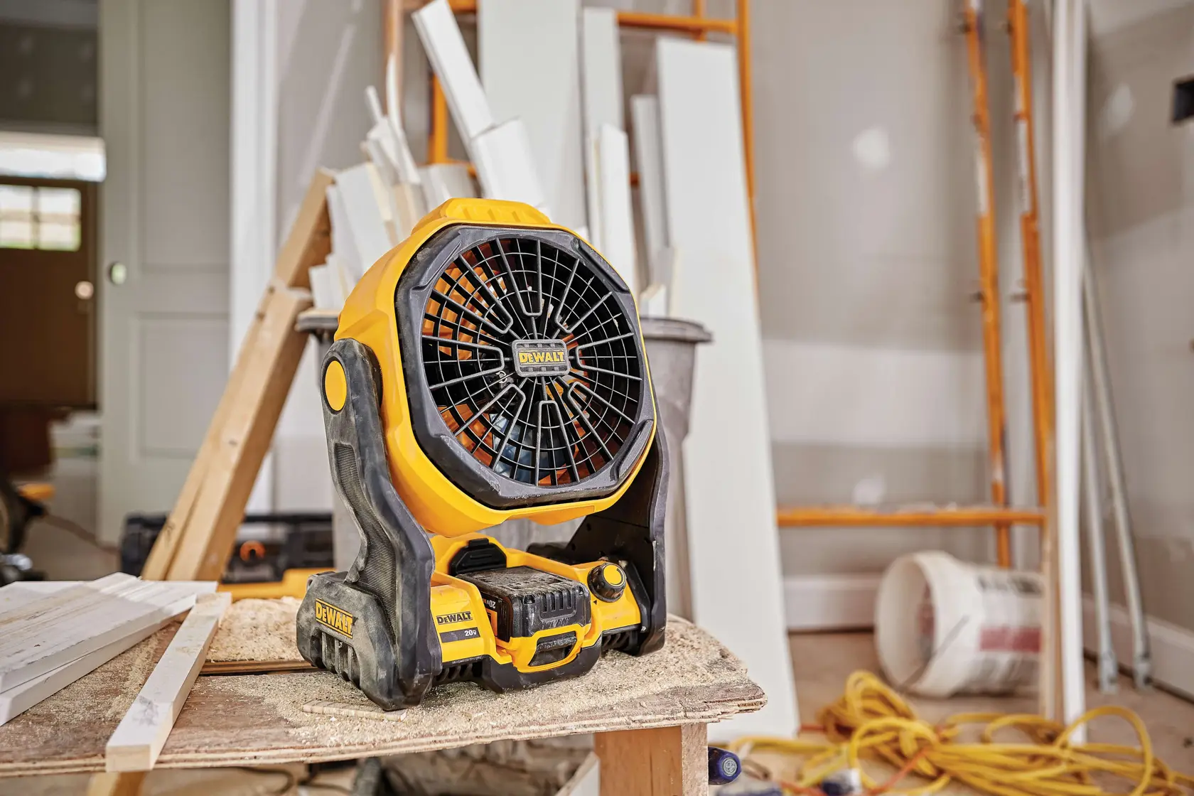 A portable DEWALT cordless fan placed on a wooden workbench in a construction or renovation setting, surrounded by tools, lumber, and equipment.