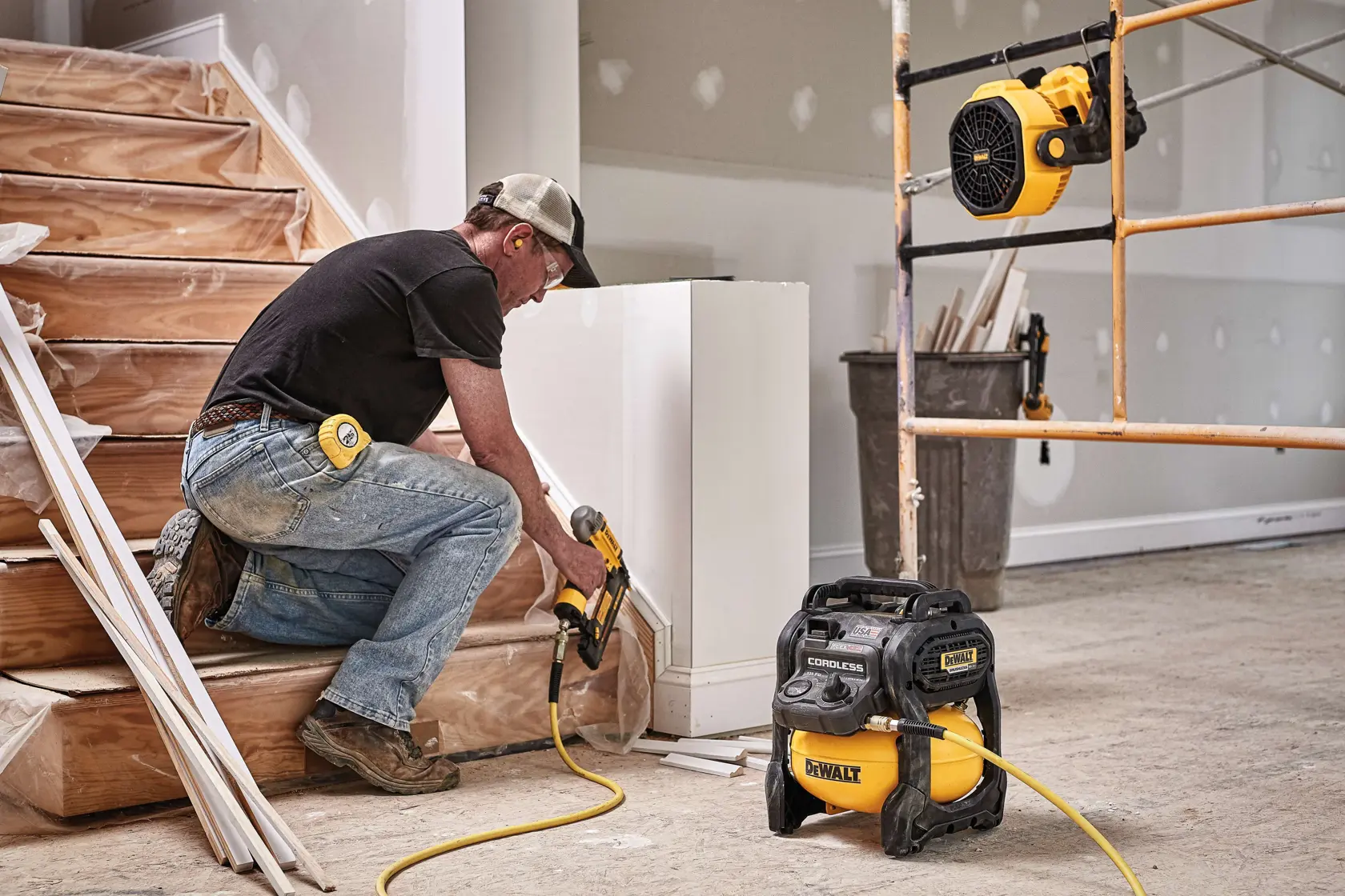 A person using a DEWALT cordless air compressor (model DCE511C1) connected to a pneumatic tool while working on stairs in a construction or renovation setting. Other DEWALT equipment and tools are visible in the background.