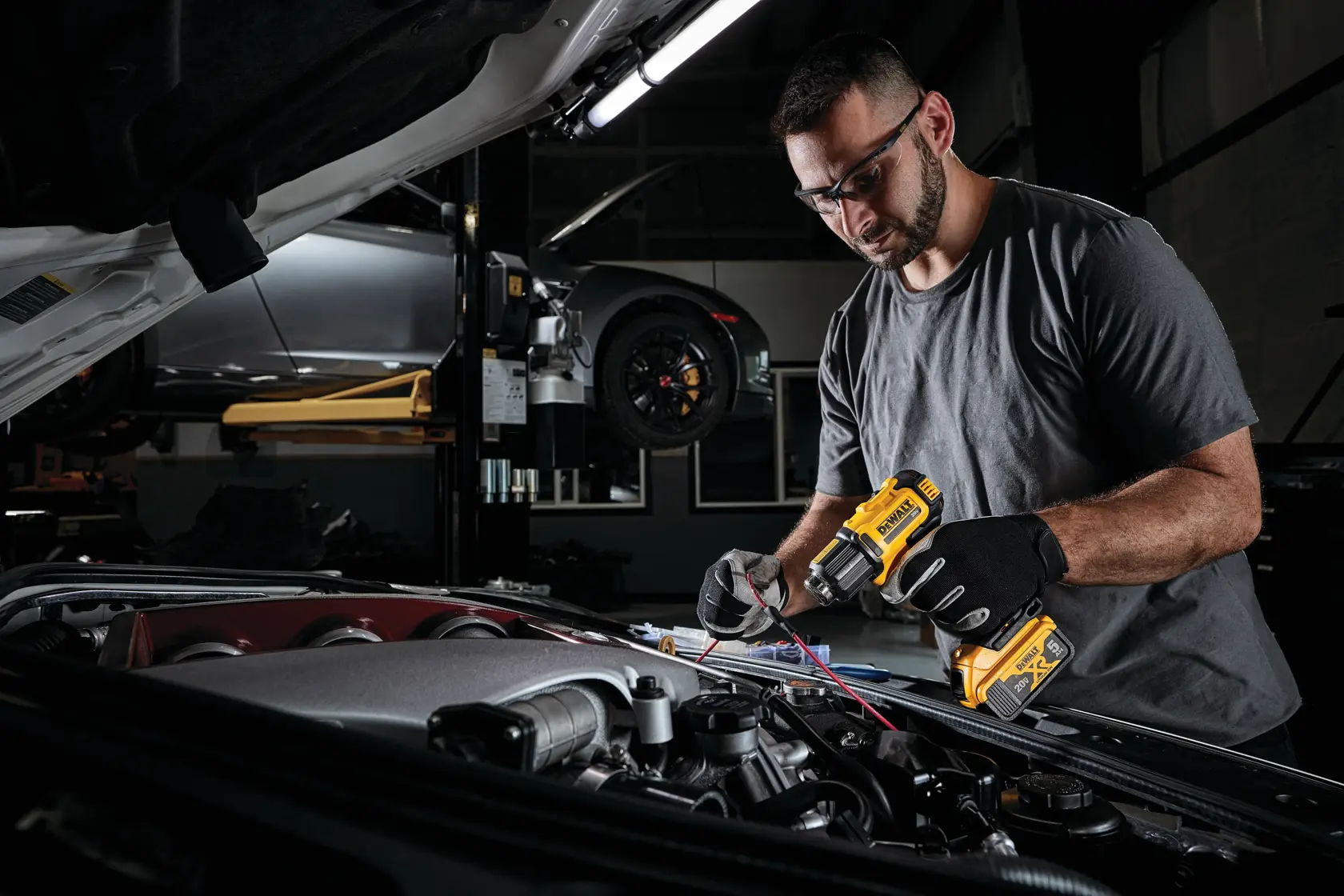A person wearing gloves is using a DEWALT cordless heat gun (DCE530B) to work on wiring under the hood of a car in a garage.